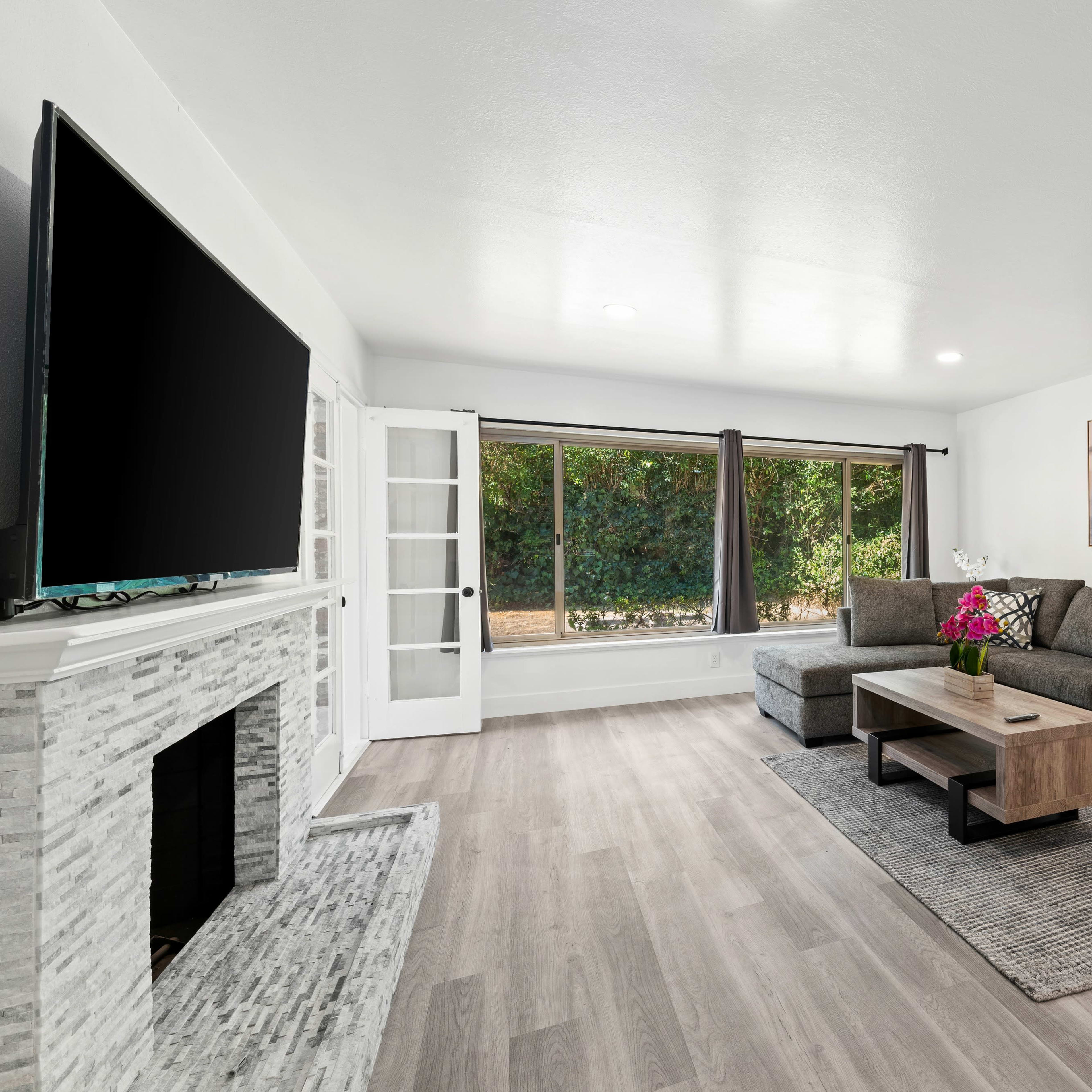A modern living room with a gray sectional sofa, a wooden coffee table, and a large TV mounted on the wall, alongside a window offering a view of greenery outside.