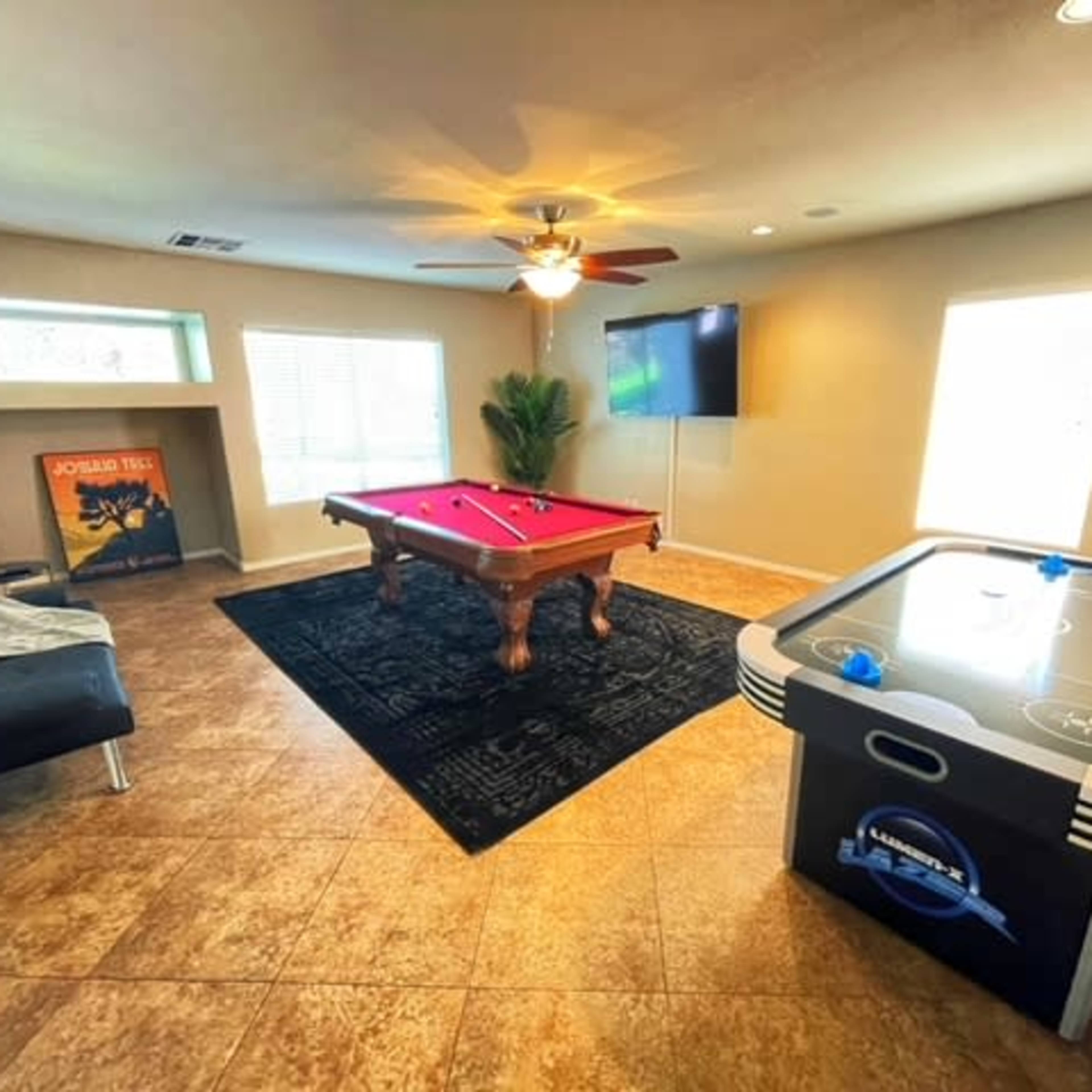 A game room featuring a billiard table, an air hockey table, and a seating area with a black sofa, illuminated by natural light from windows.