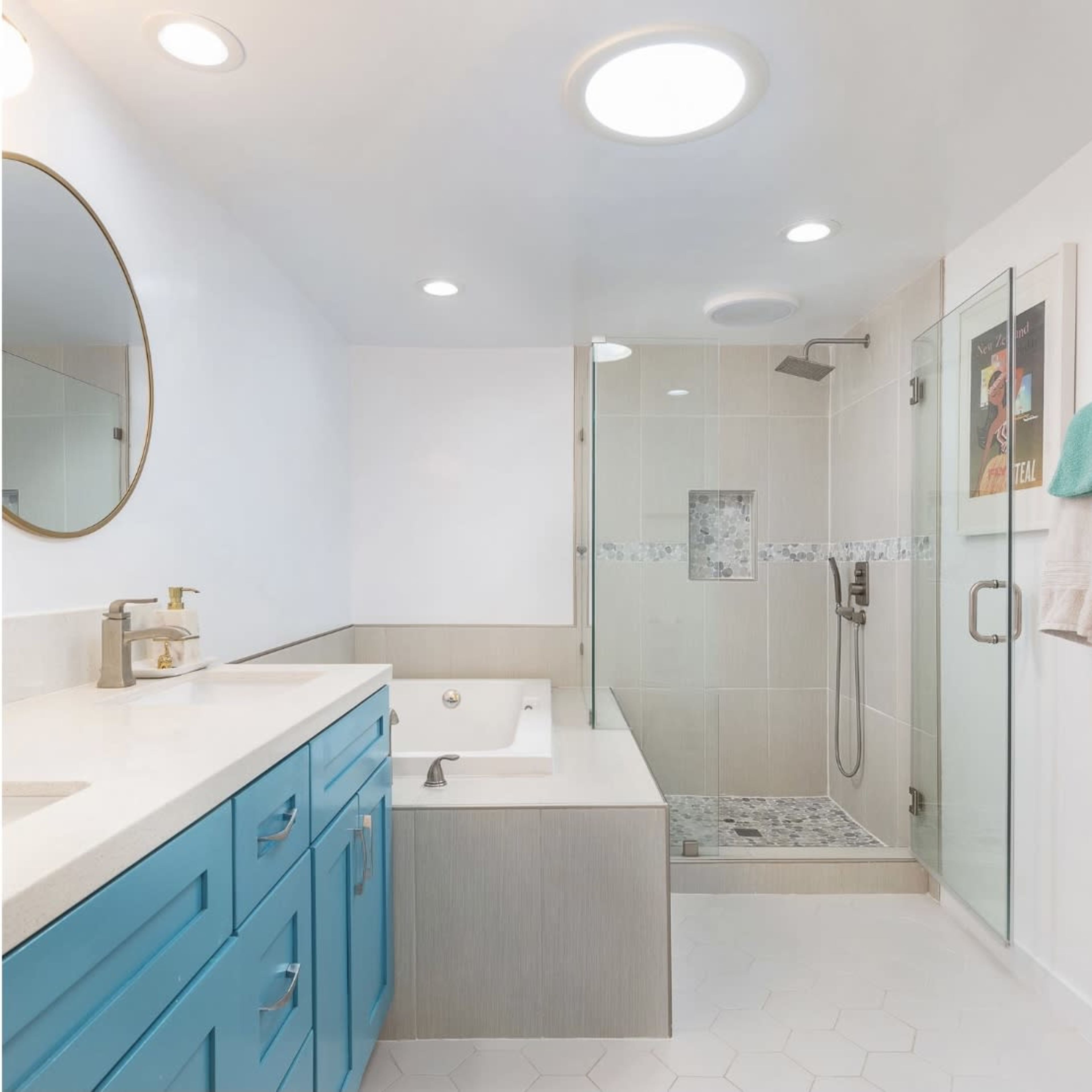 A modern bathroom featuring a blue vanity, a glass shower enclosure, and a freestanding bathtub, with ample white lighting and a round mirror.