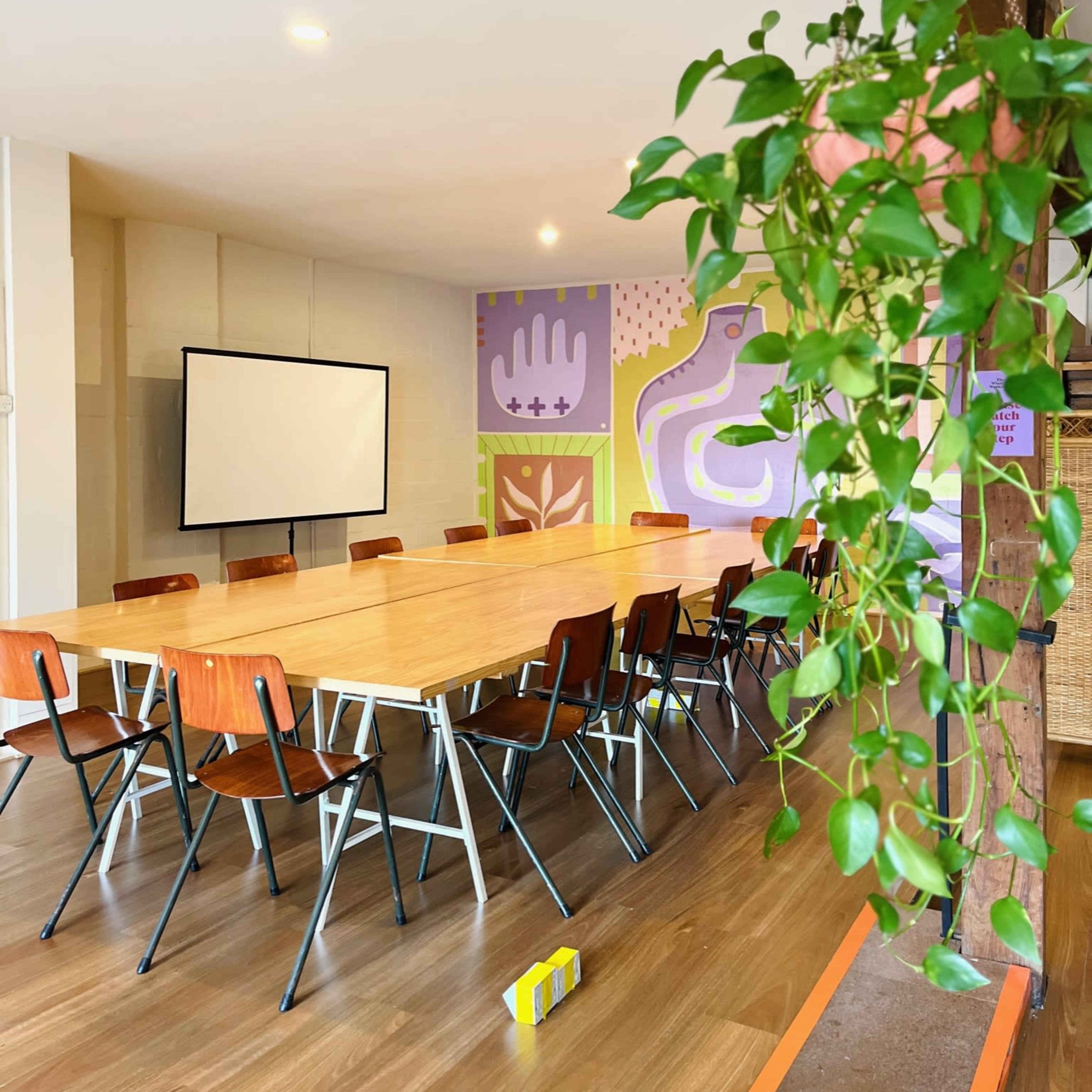 A spacious meeting room with a long wooden table surrounded by chairs, featuring colorful wall murals and a projector screen at the front.