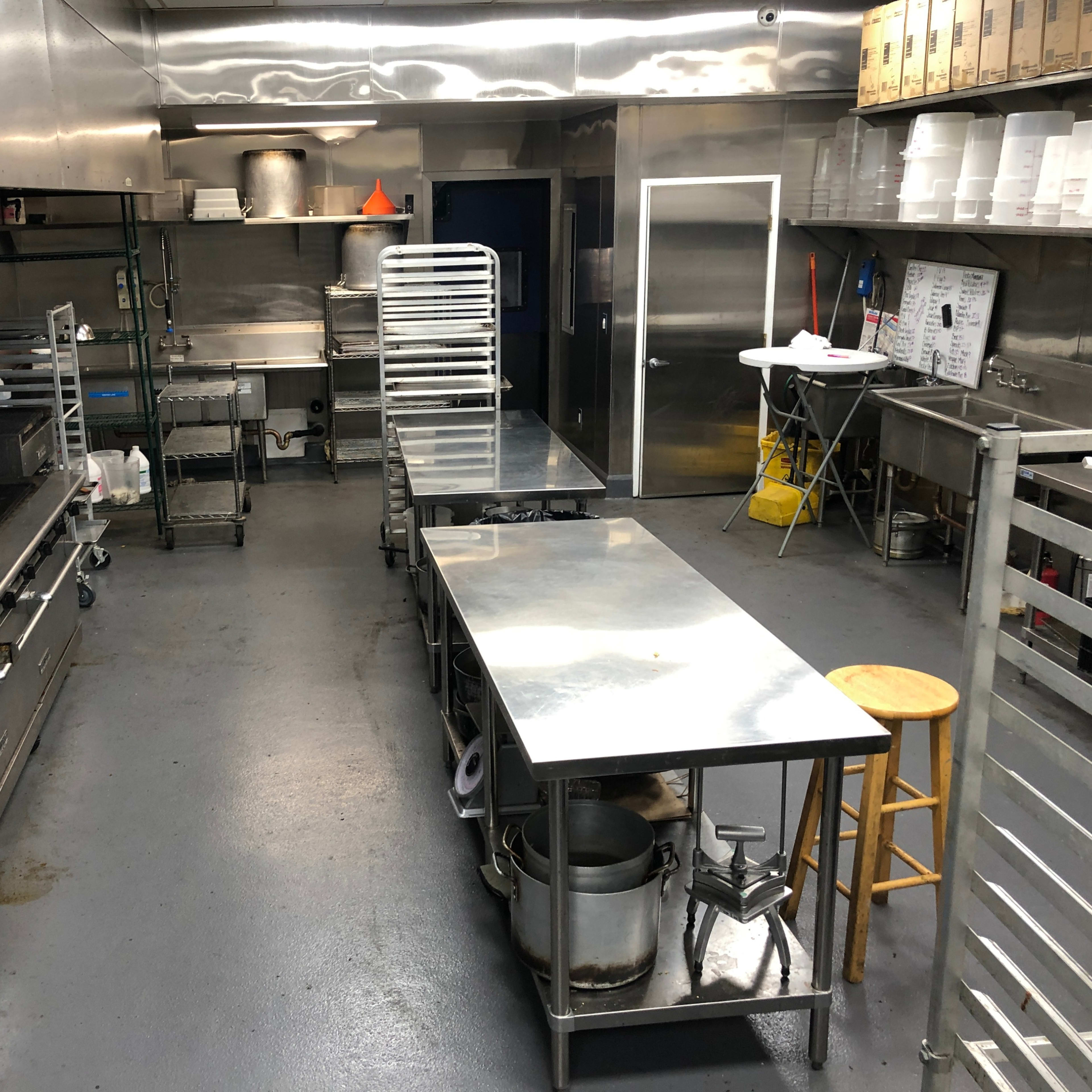A stainless steel commercial kitchen with multiple worktables, cooking equipment, and shelves.