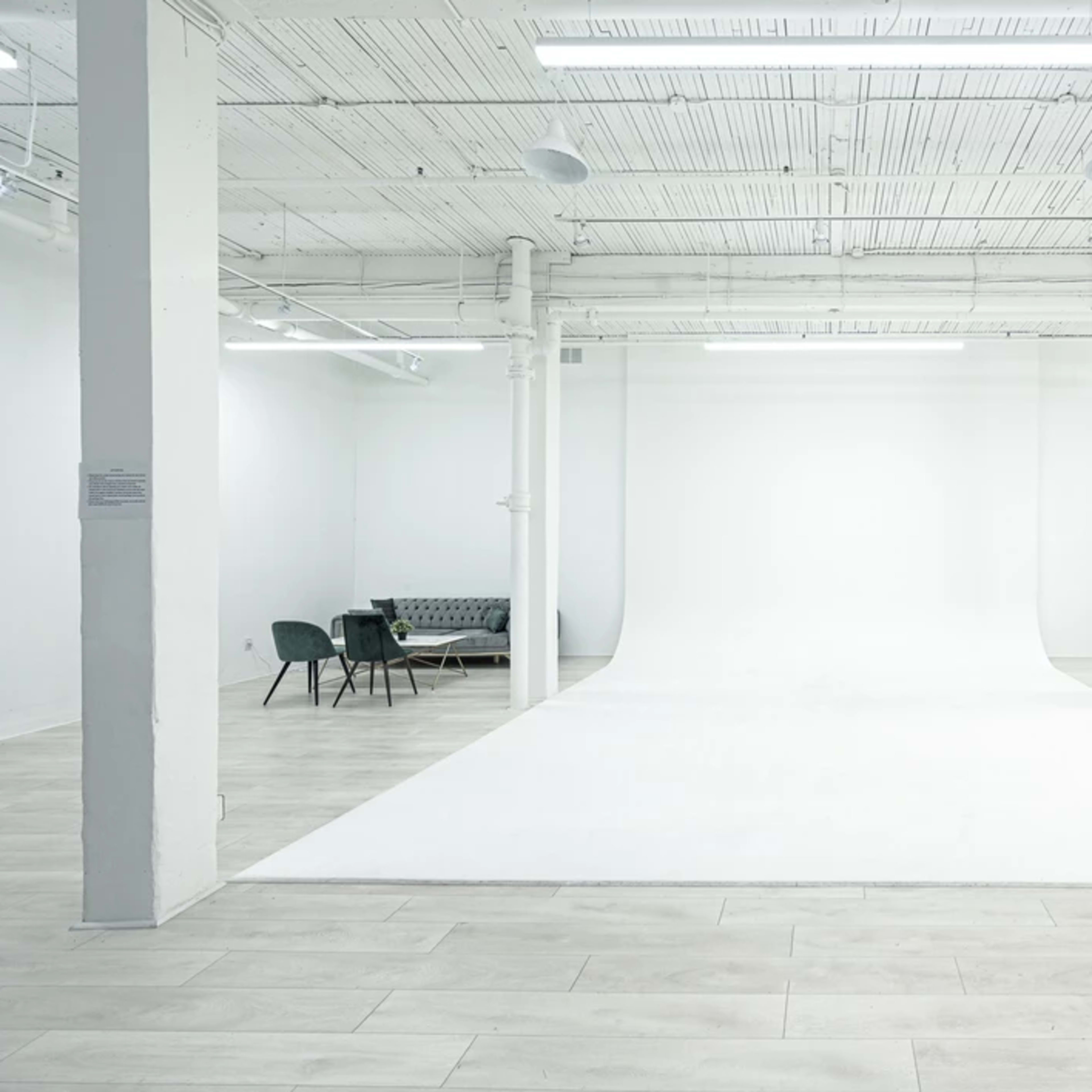 A spacious, modern studio with a white backdrop, wooden flooring, and a seating area consisting of chairs and a couch.