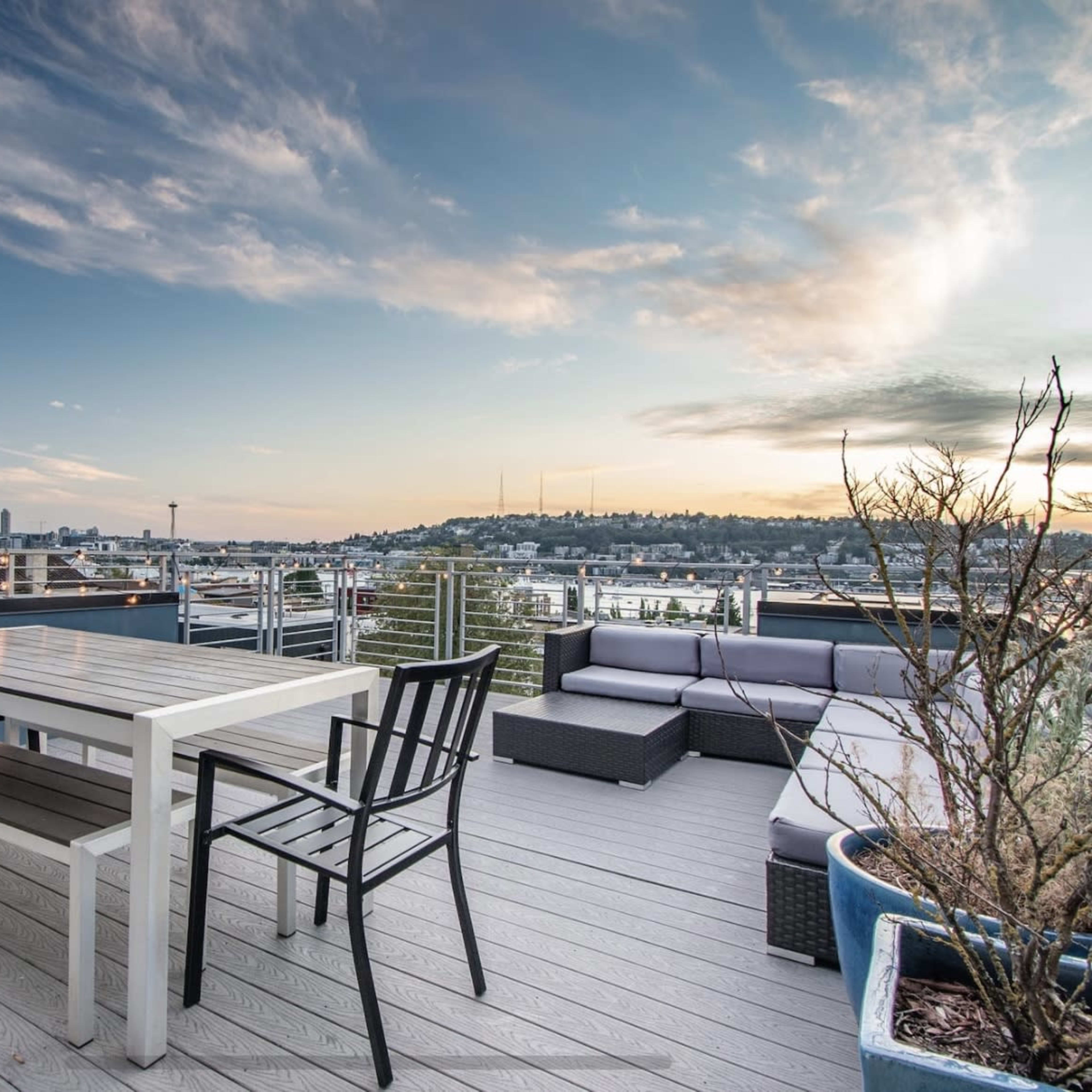 A rooftop terrace features a white dining table and black chairs, overlooking a city skyline and dotted with potted plants.