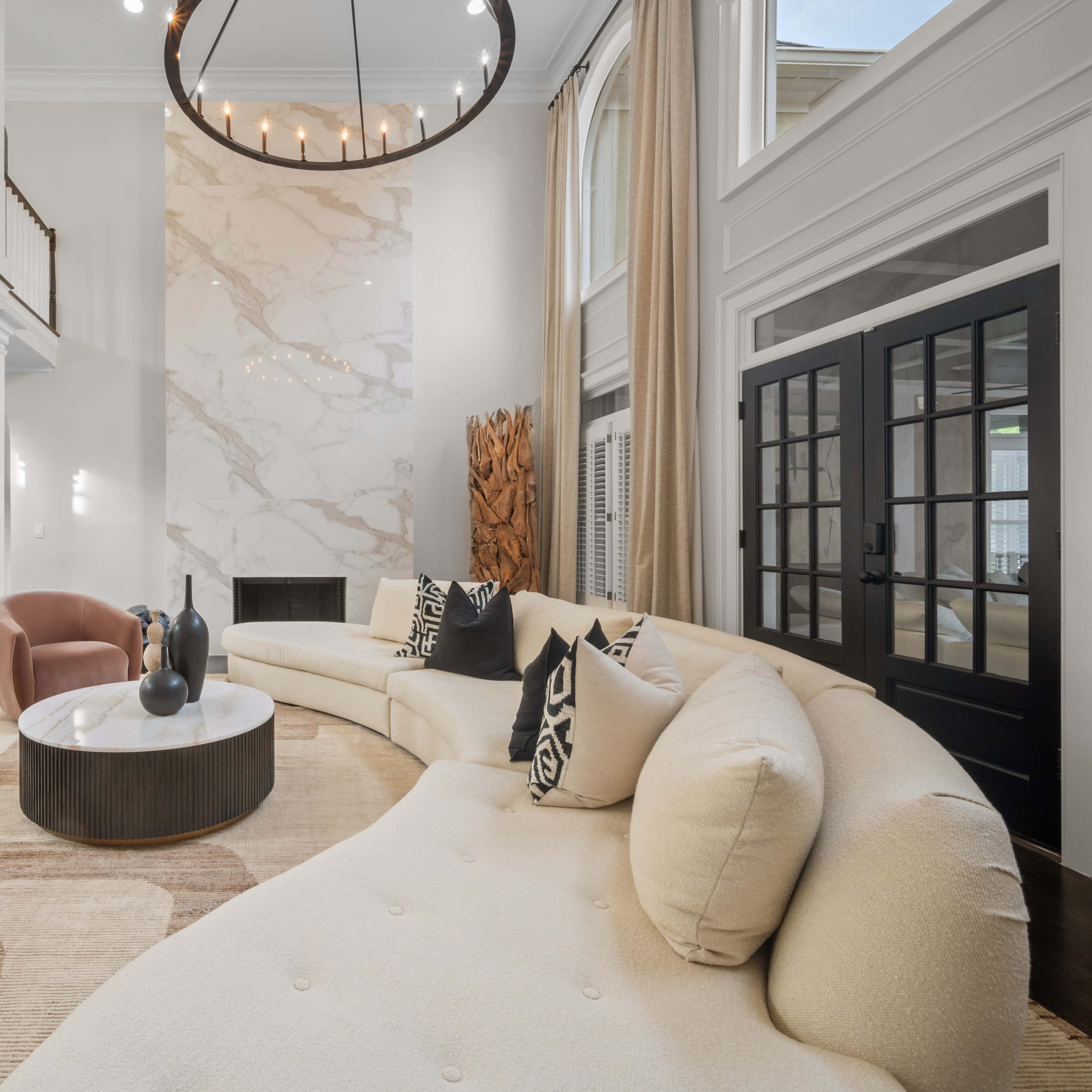 A large, modern living room features a curved sofa, a round coffee table, and a light-colored marble accent wall under a circular chandelier.