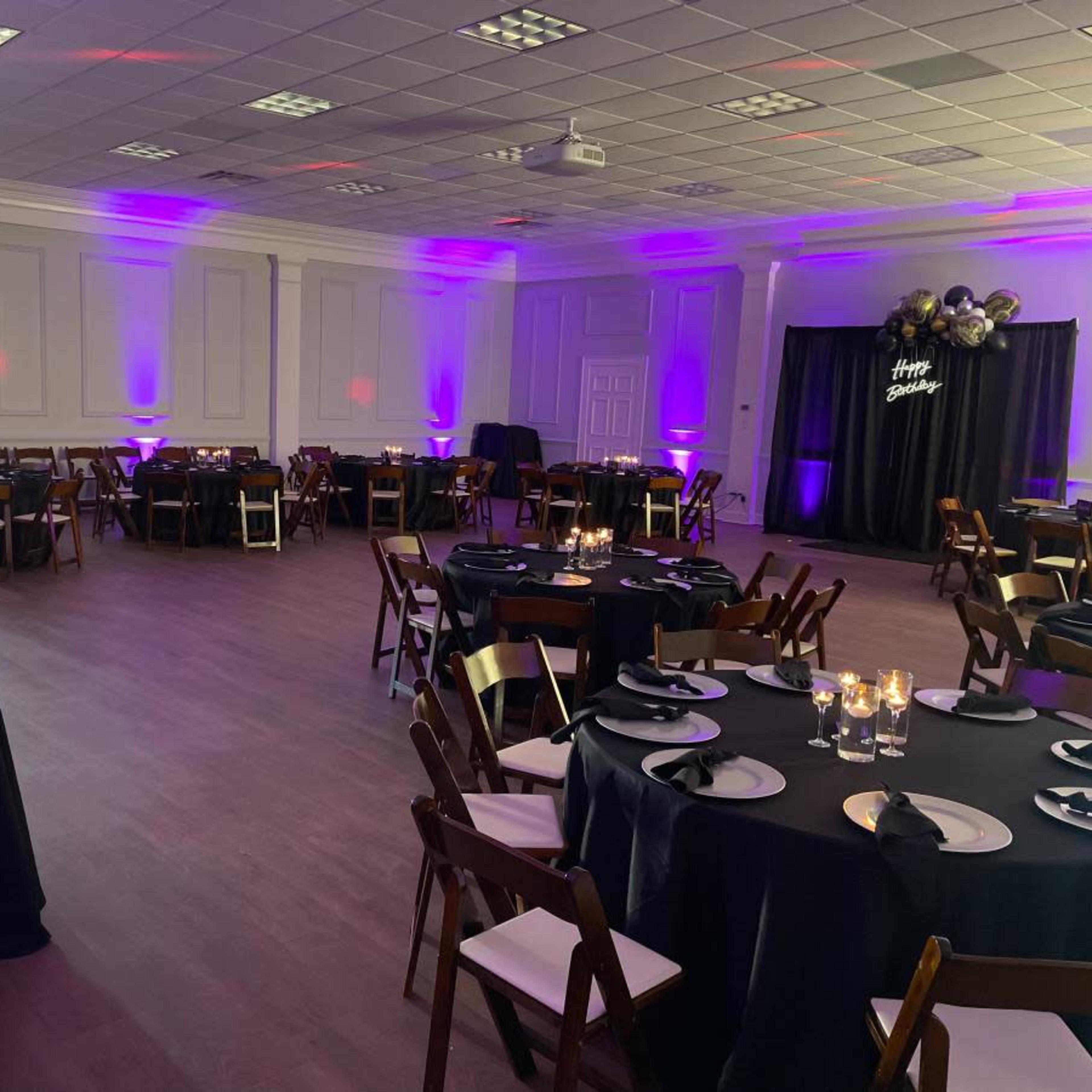 The interior of a banquet hall features neatly arranged round tables with black tablecloths, wooden chairs, and purple lighting, set up for an event.
