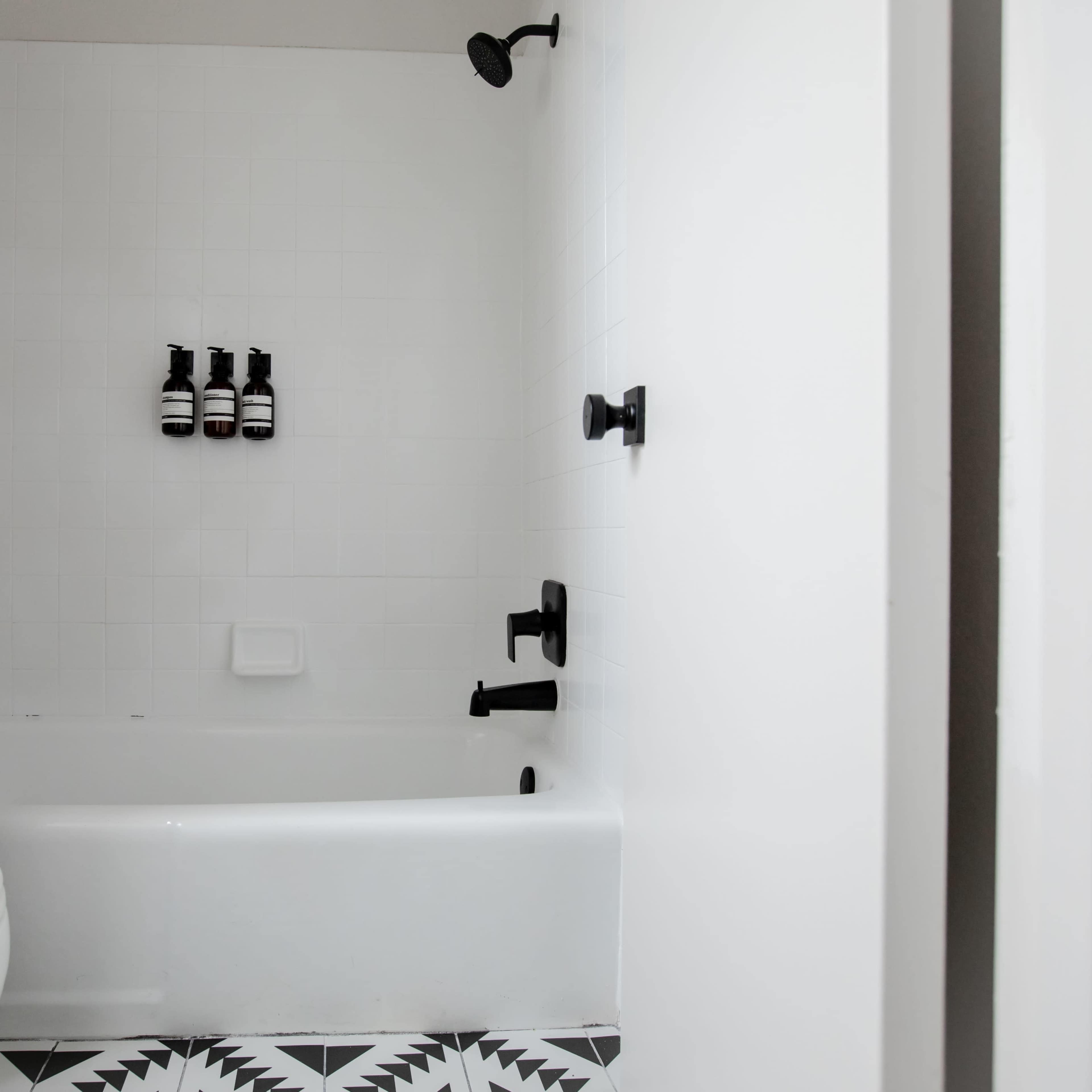 A bathroom with a bathtub, a showerhead, and three black bottles on the wall, featuring a patterned floor and a curtain beside the bathtub.