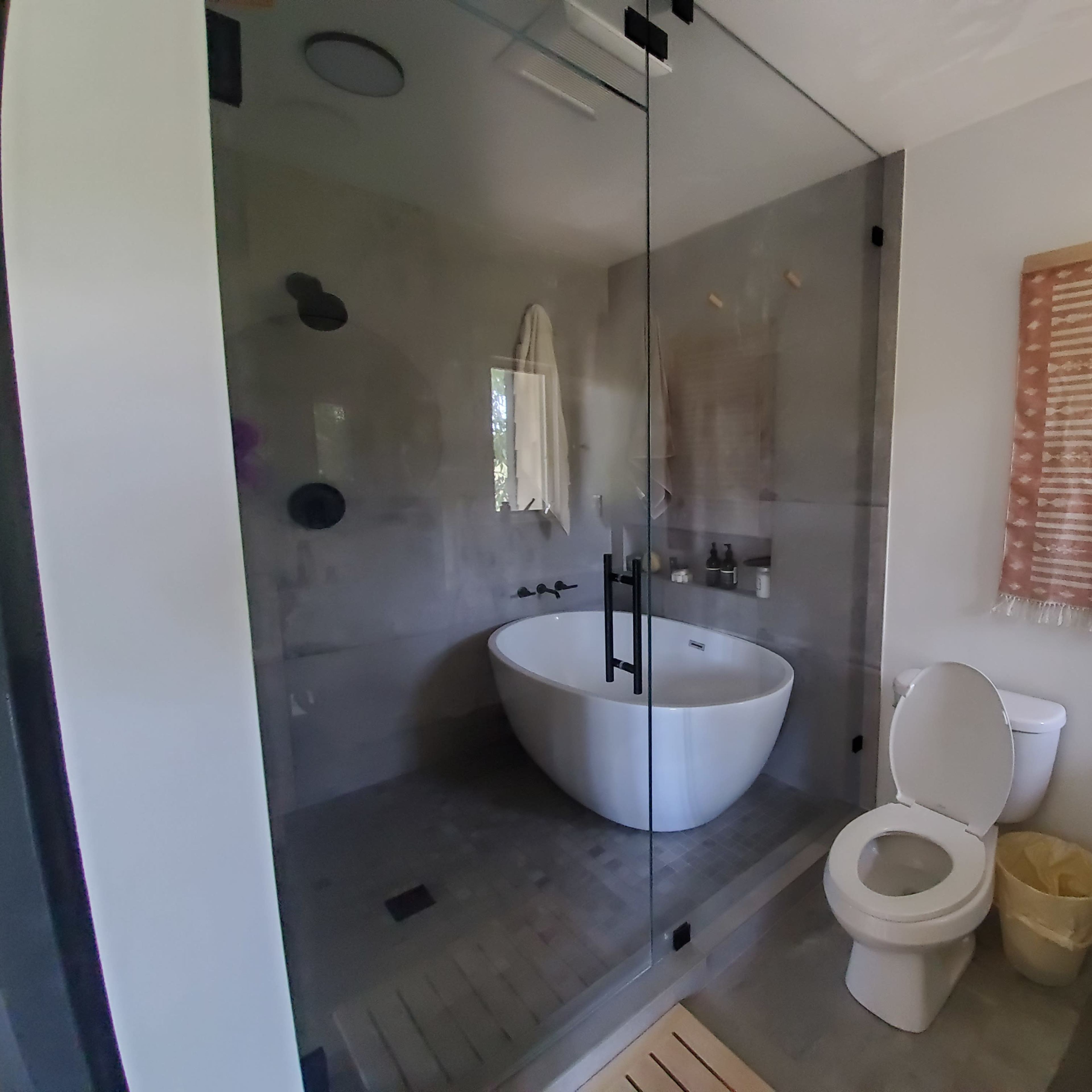 A modern bathroom featuring a glass shower enclosure, a freestanding bathtub, a toilet, and a woven wall hanging.