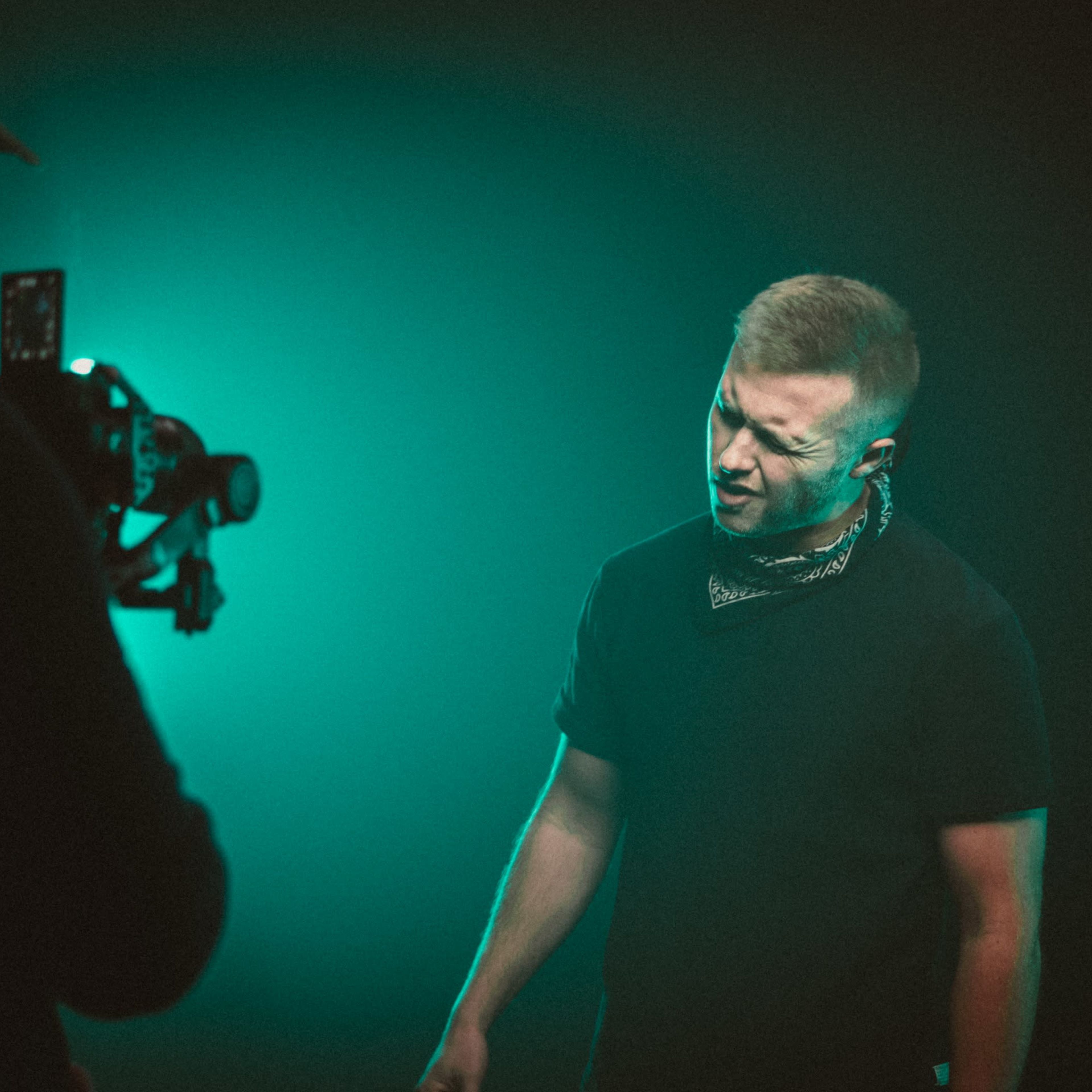 A man in a black shirt and bandana performs in front of a camera with blue-green lighting in a studio setting.