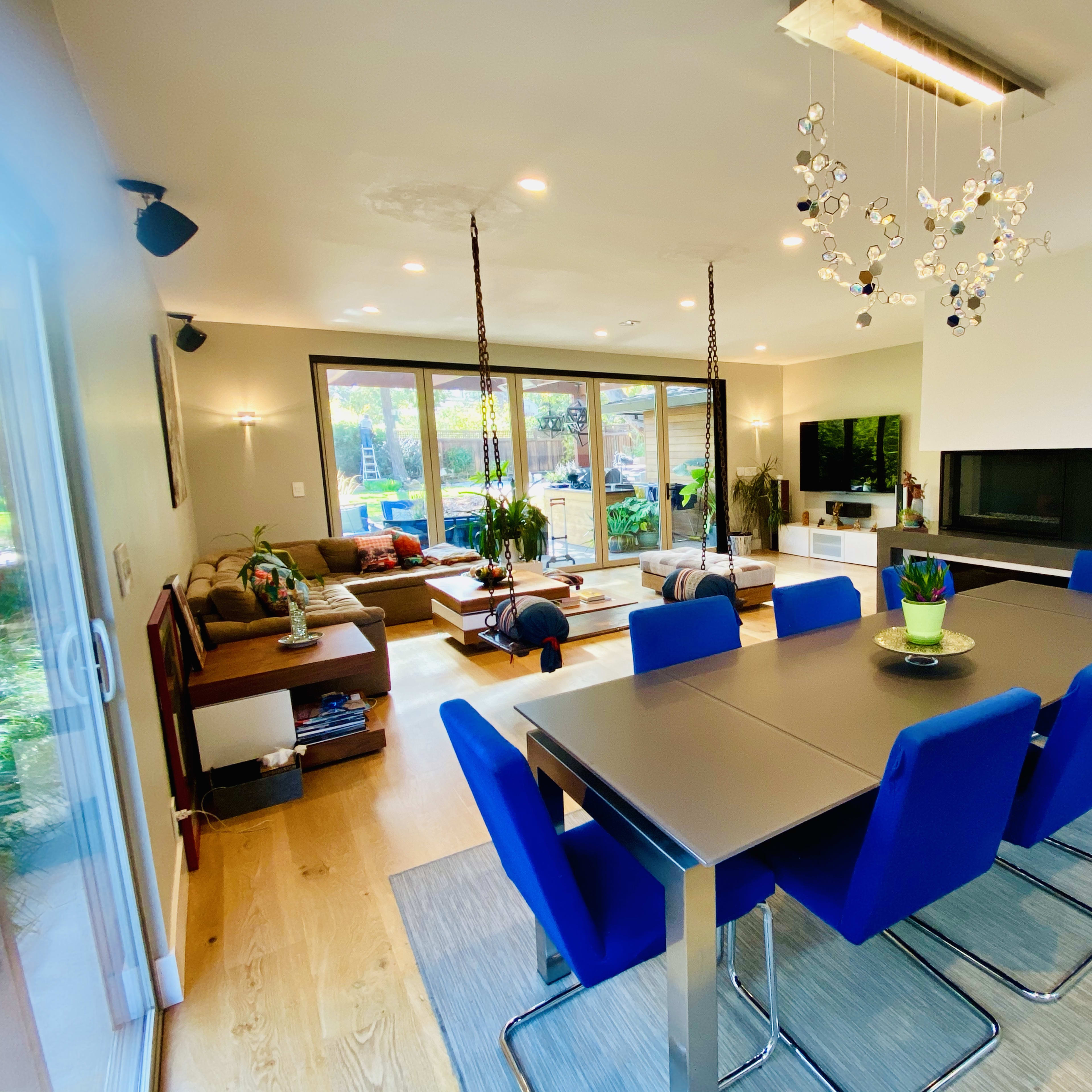 A modern dining area features a sleek table with blue chairs, a chandelier, and large windows overlooking a backyard with greenery.