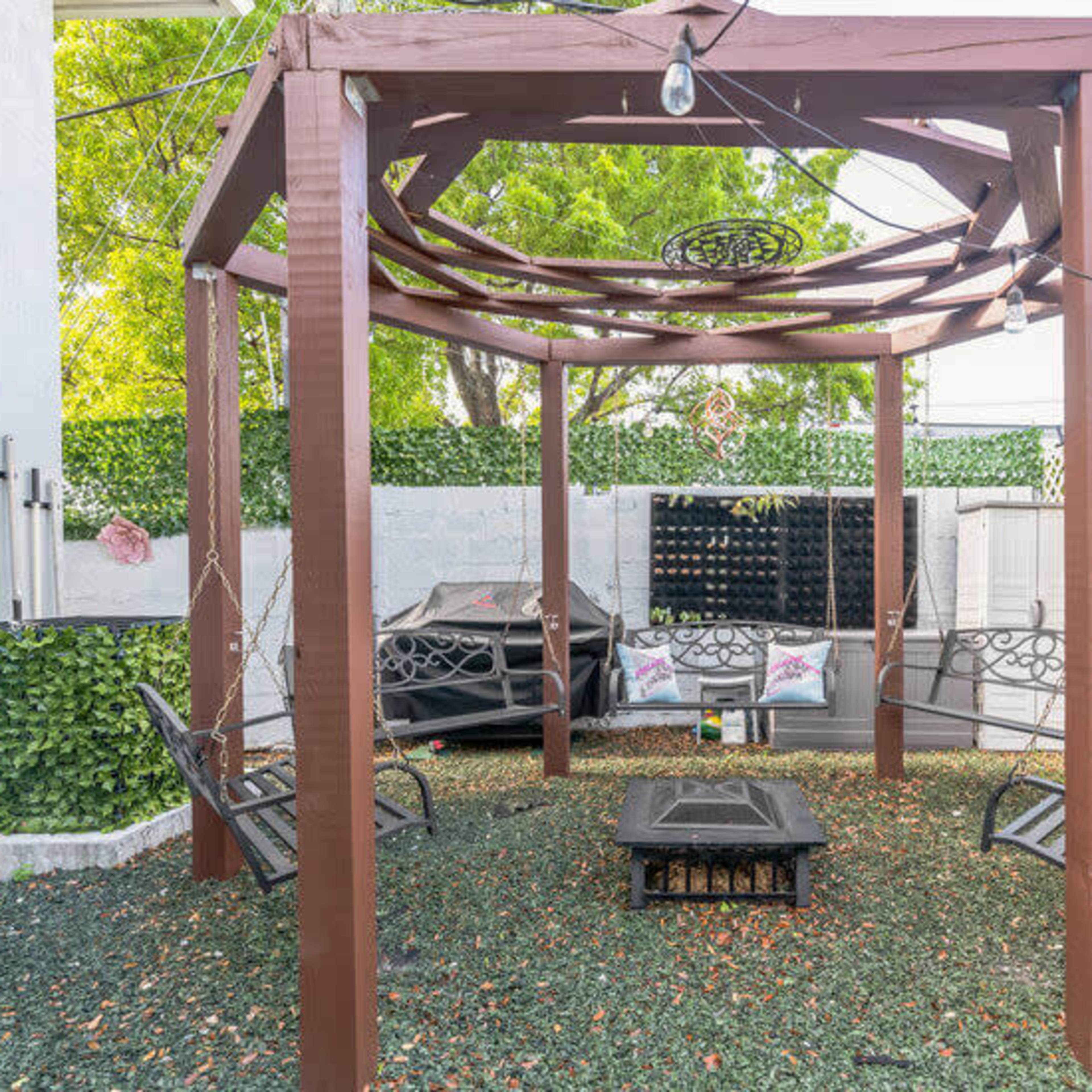 A wooden gazebo with swing seating is surrounded by greenery and outdoor furnishings in a spacious outdoor area.
