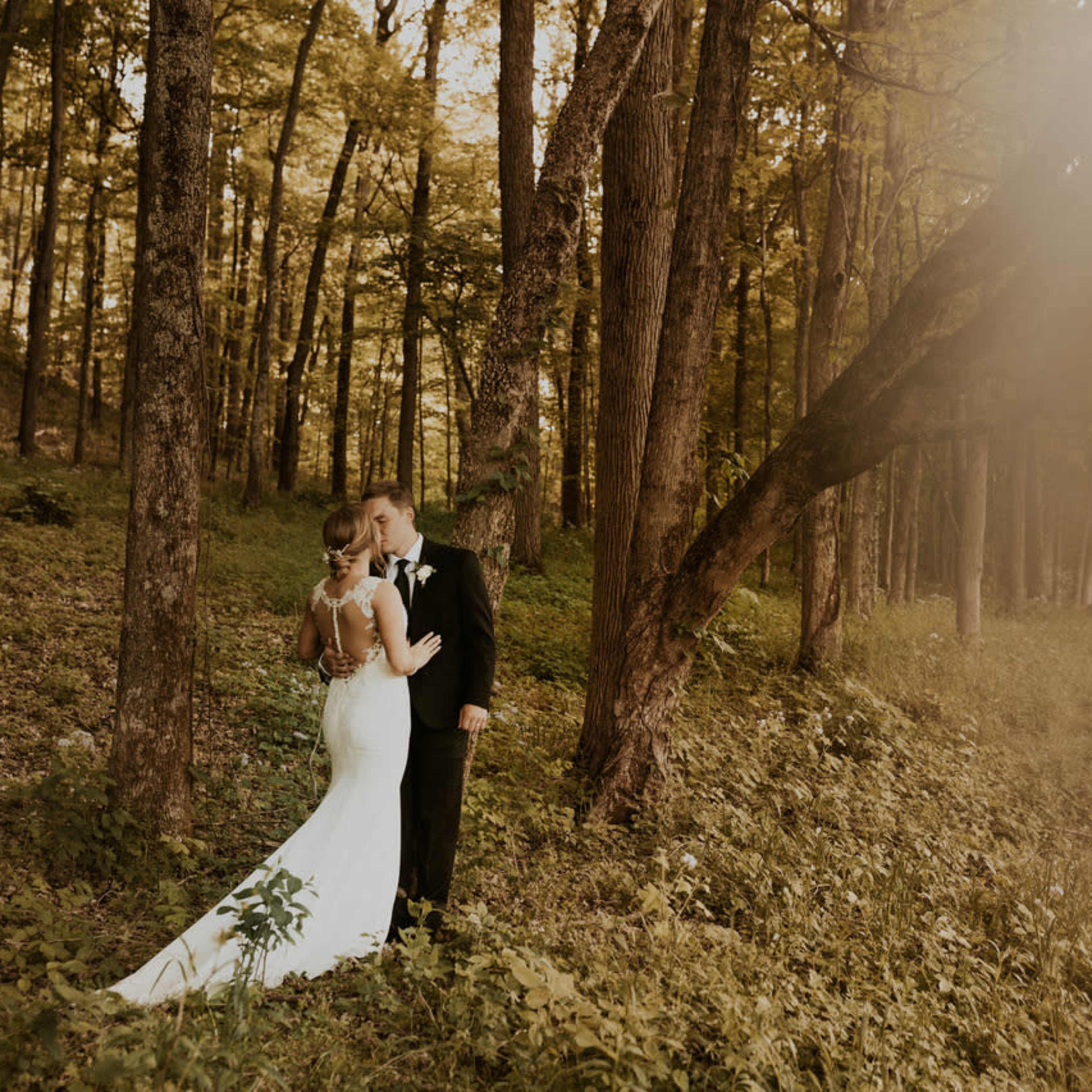 A couple stands embraced in a wooded area, surrounded by tall trees and greenery.