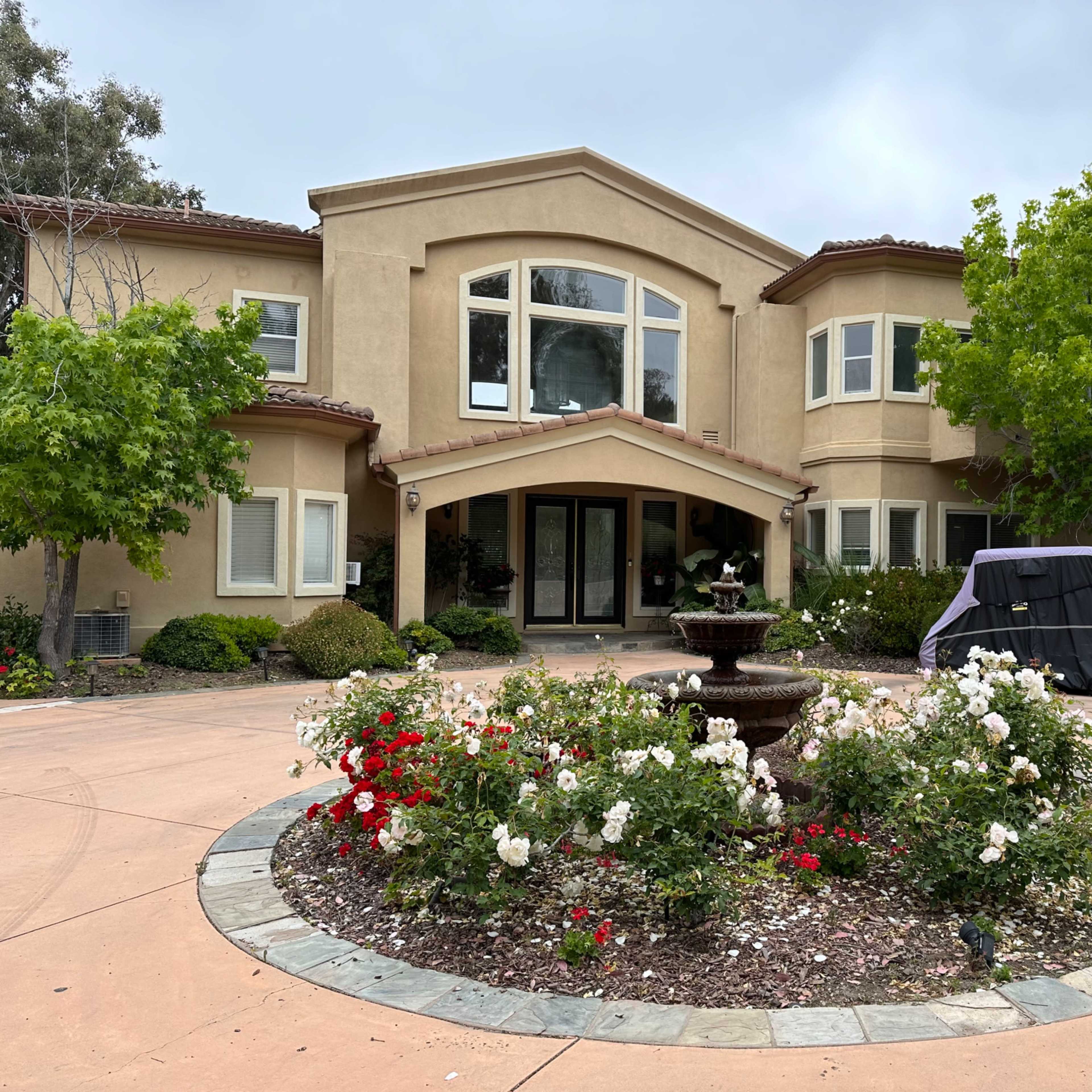 A large, two-story house with a circular garden and a fountain features blooming rose bushes in front.