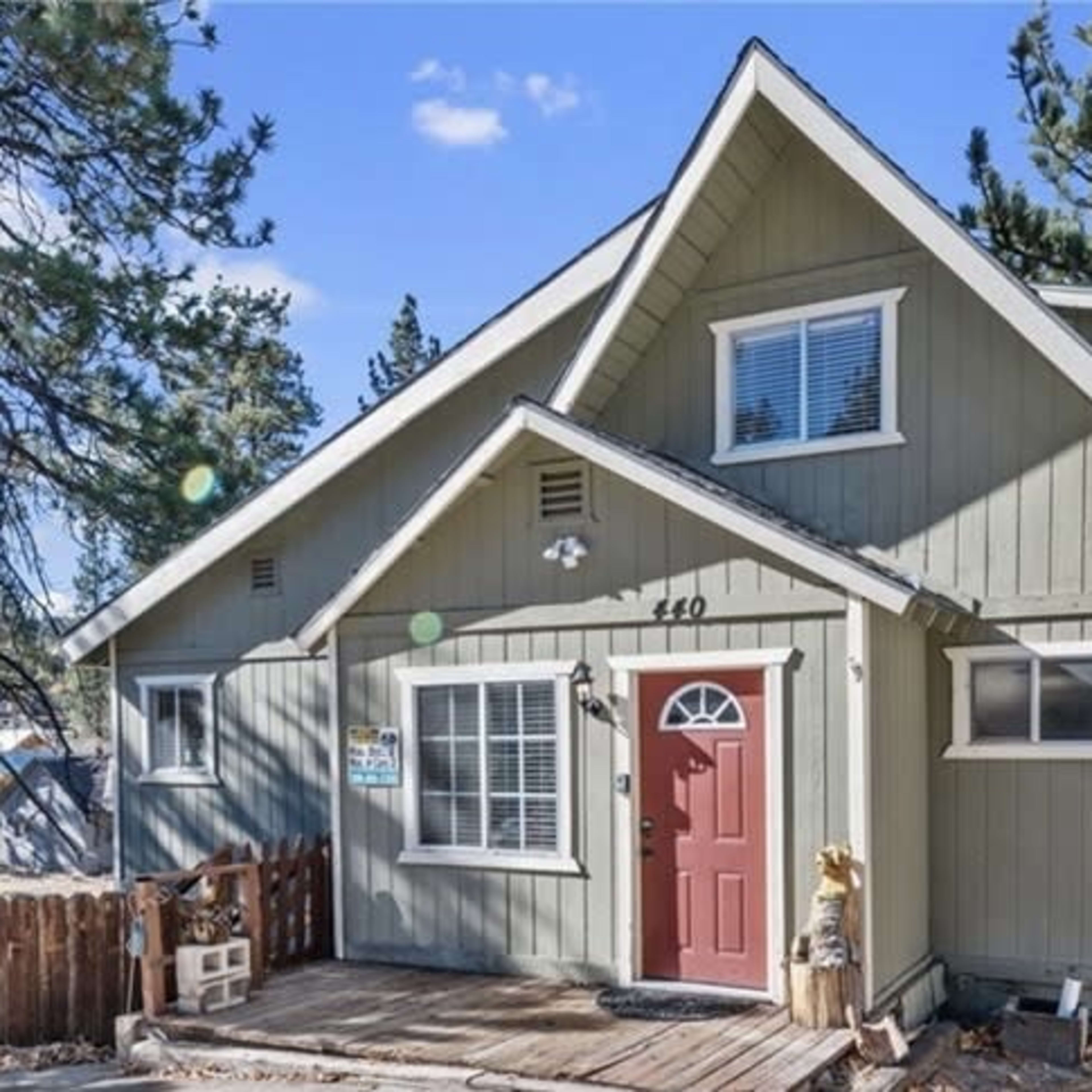 A green, two-story house with a red door and a wooden porch is surrounded by tall pine trees.