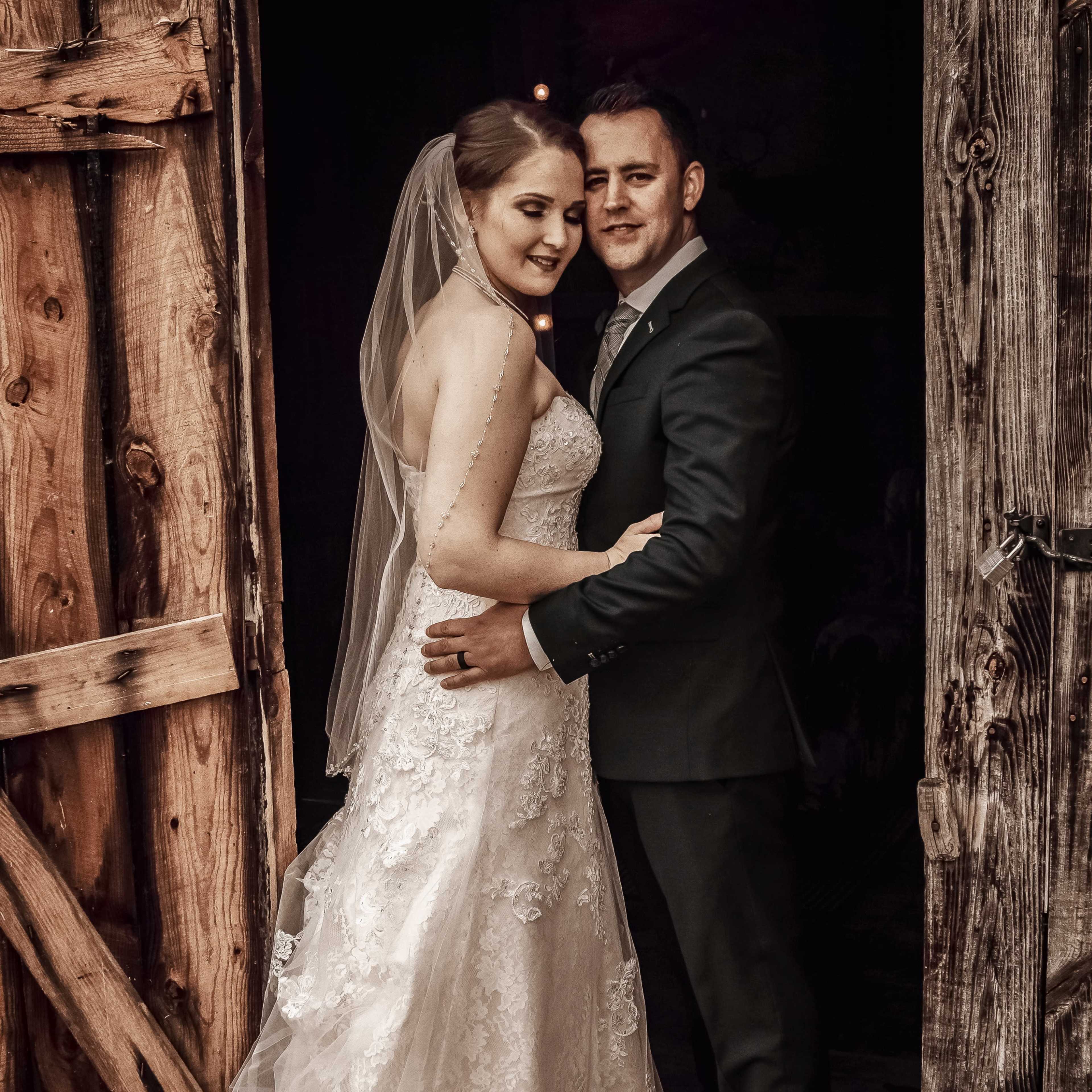 A bride in a white lace gown and a groom in a suit pose together at an entrance of a rustic wooden building.