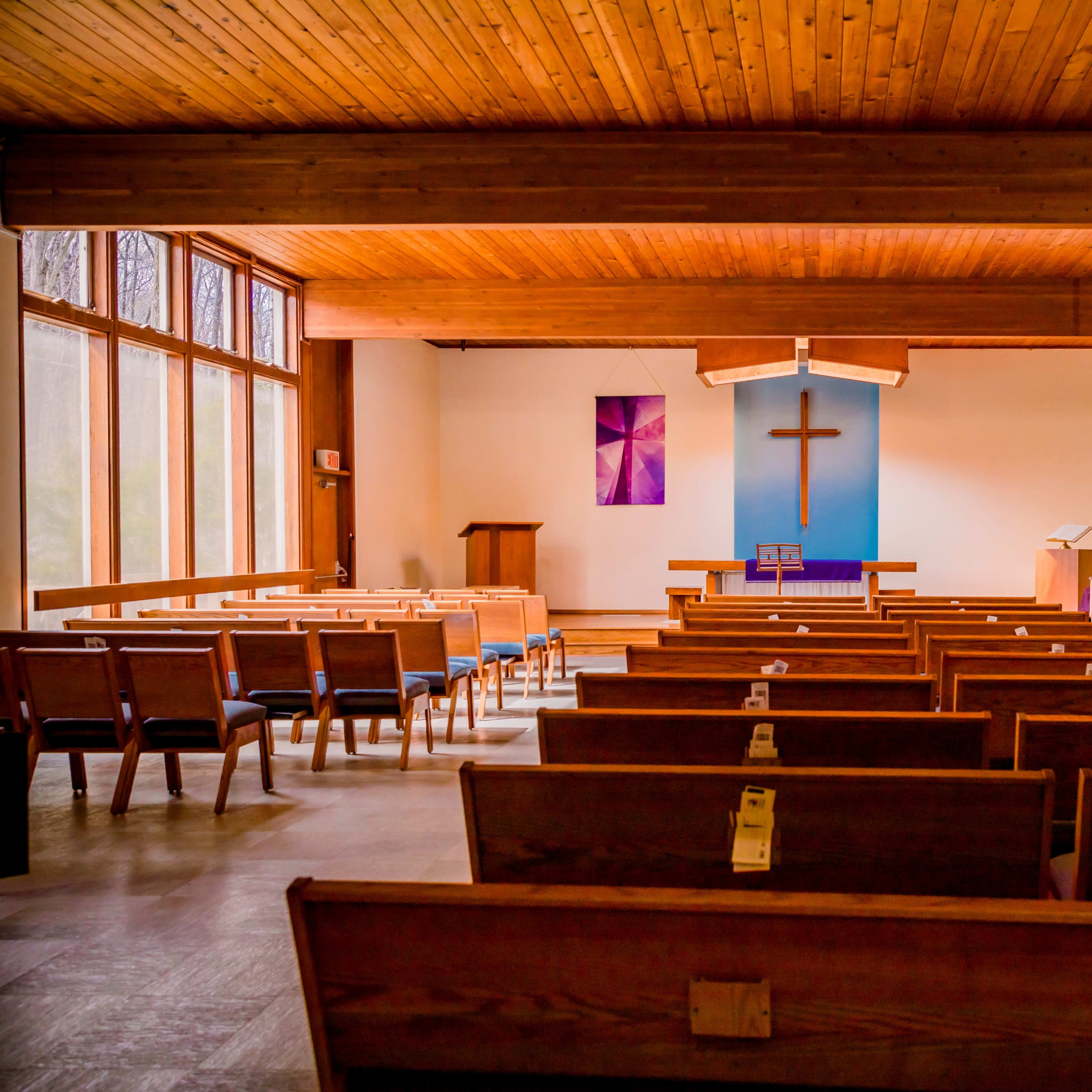 The interior of a church with wooden benches facing a blue wall featuring a large cross.