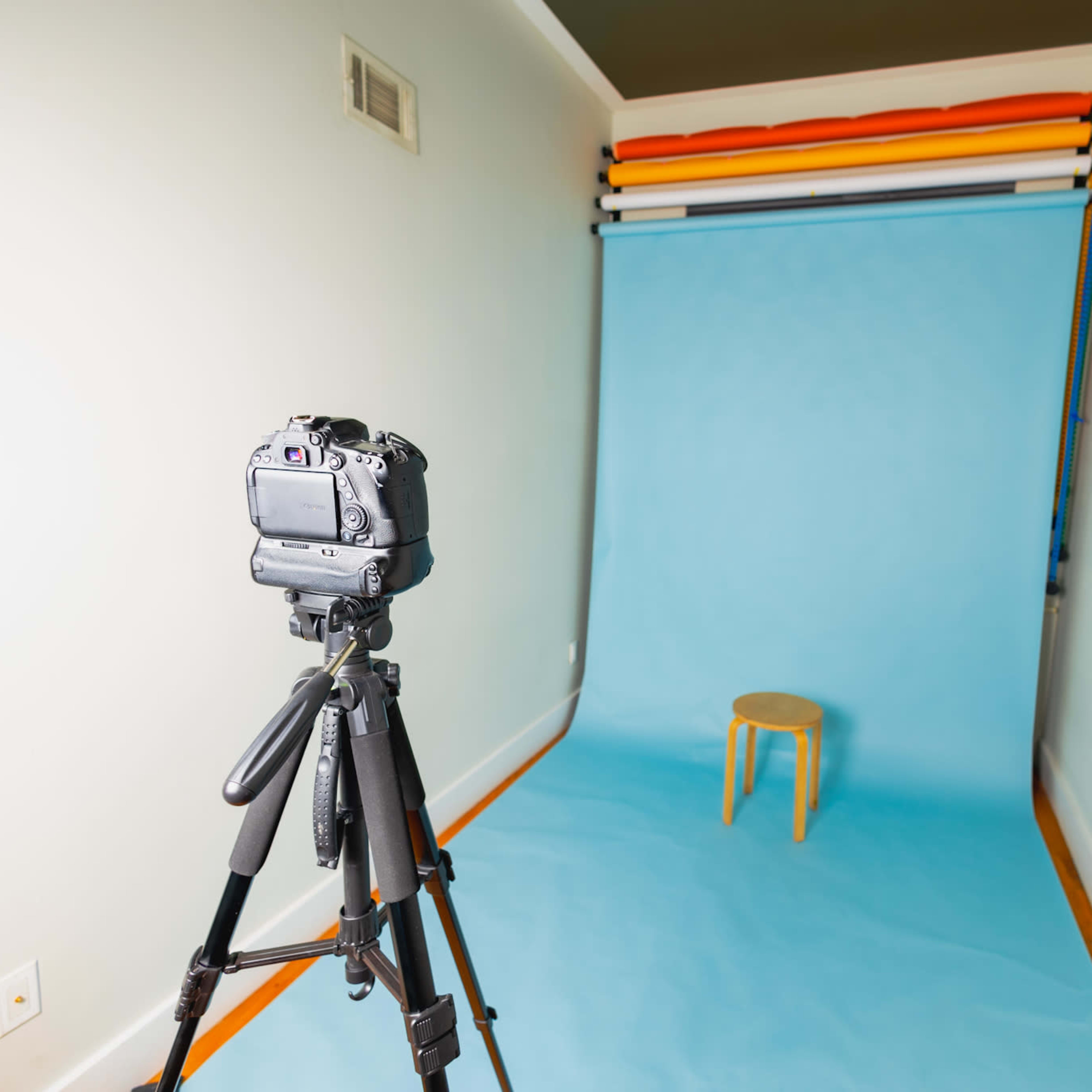 A camera on a tripod faces a plain blue backdrop with a wooden stool set in the center.