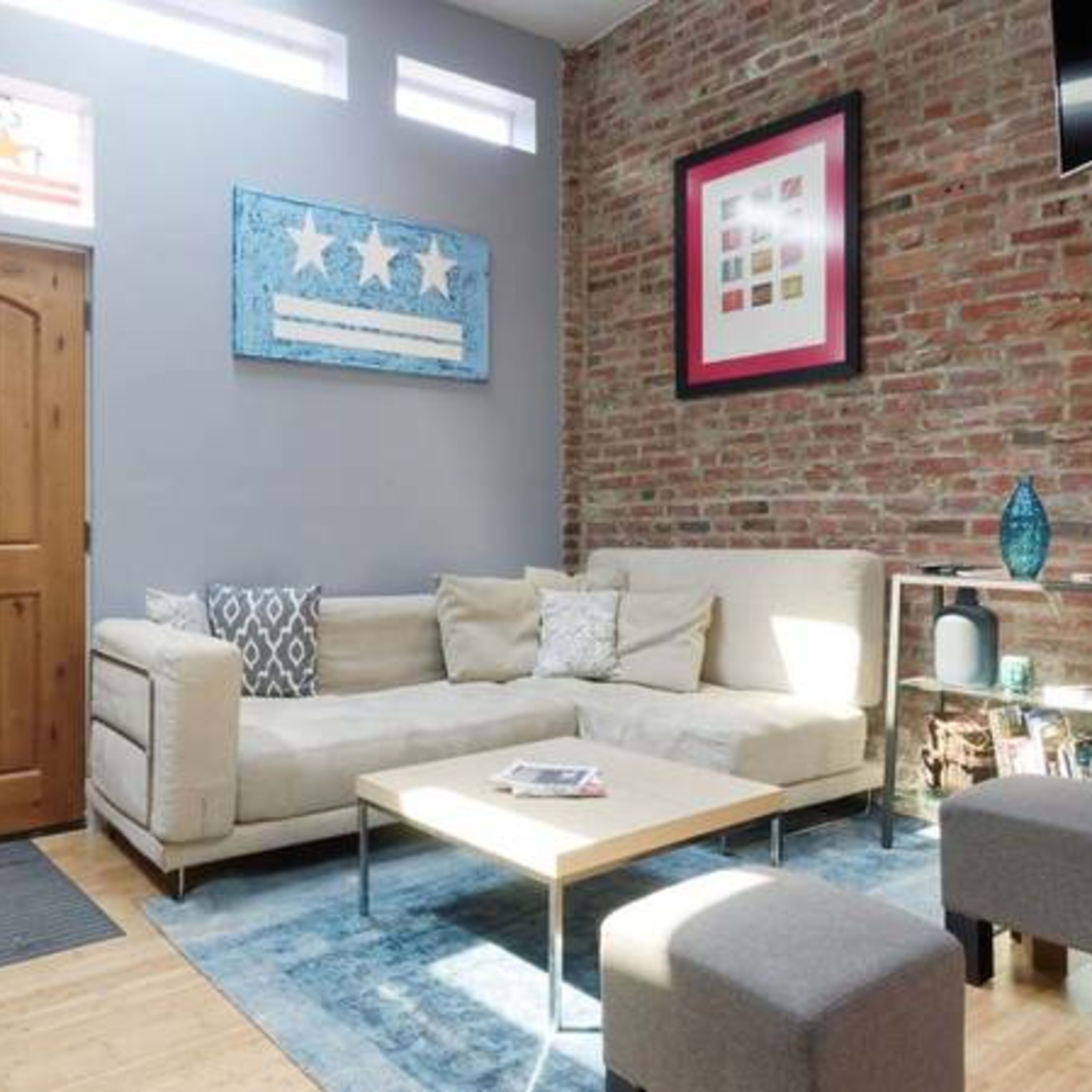 A modern living room with a beige sectional sofa, a wooden door, and a coffee table on a blue rug, framed by exposed brick walls.