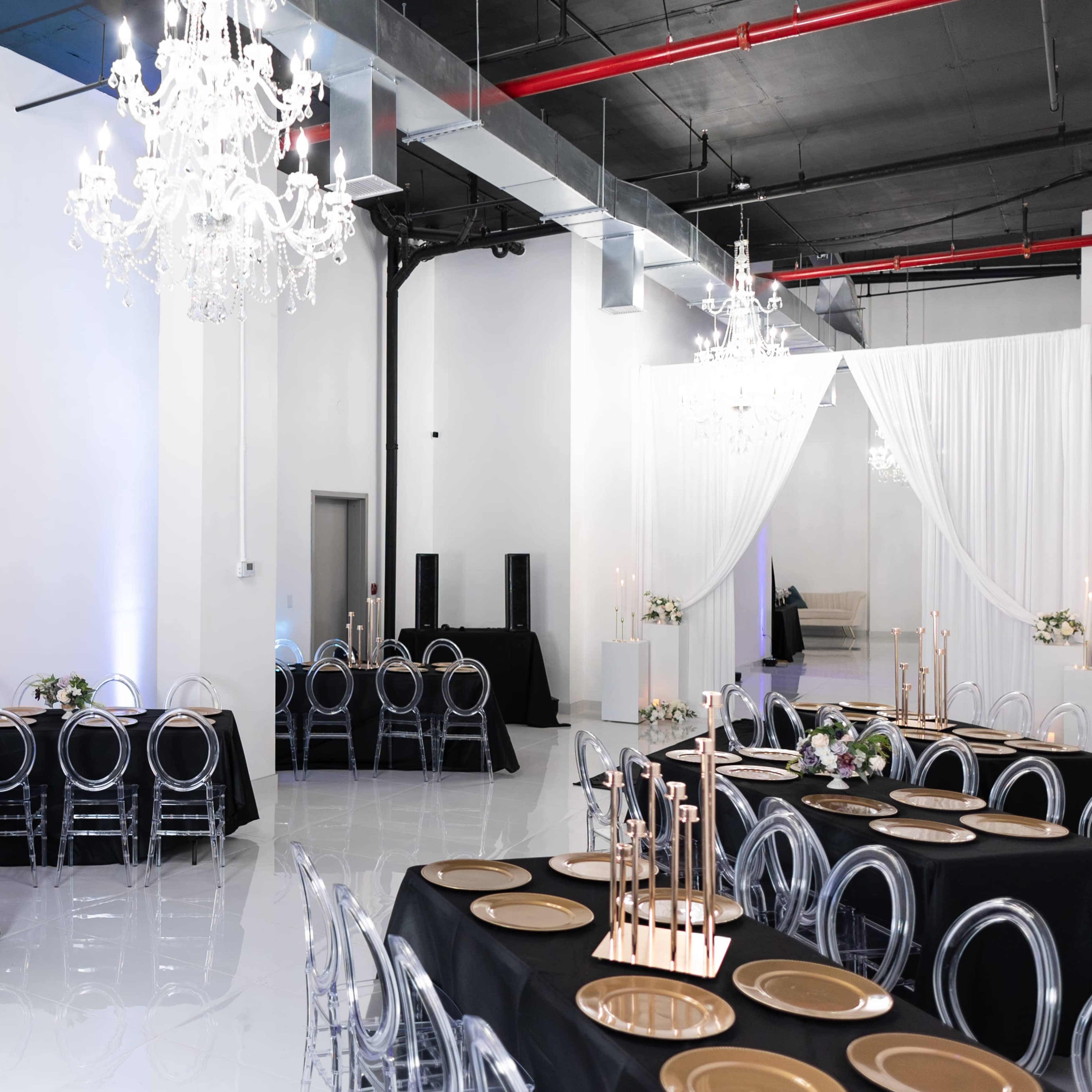 A spacious event hall features black tables set with gold chargers and illuminated by chandeliers.
