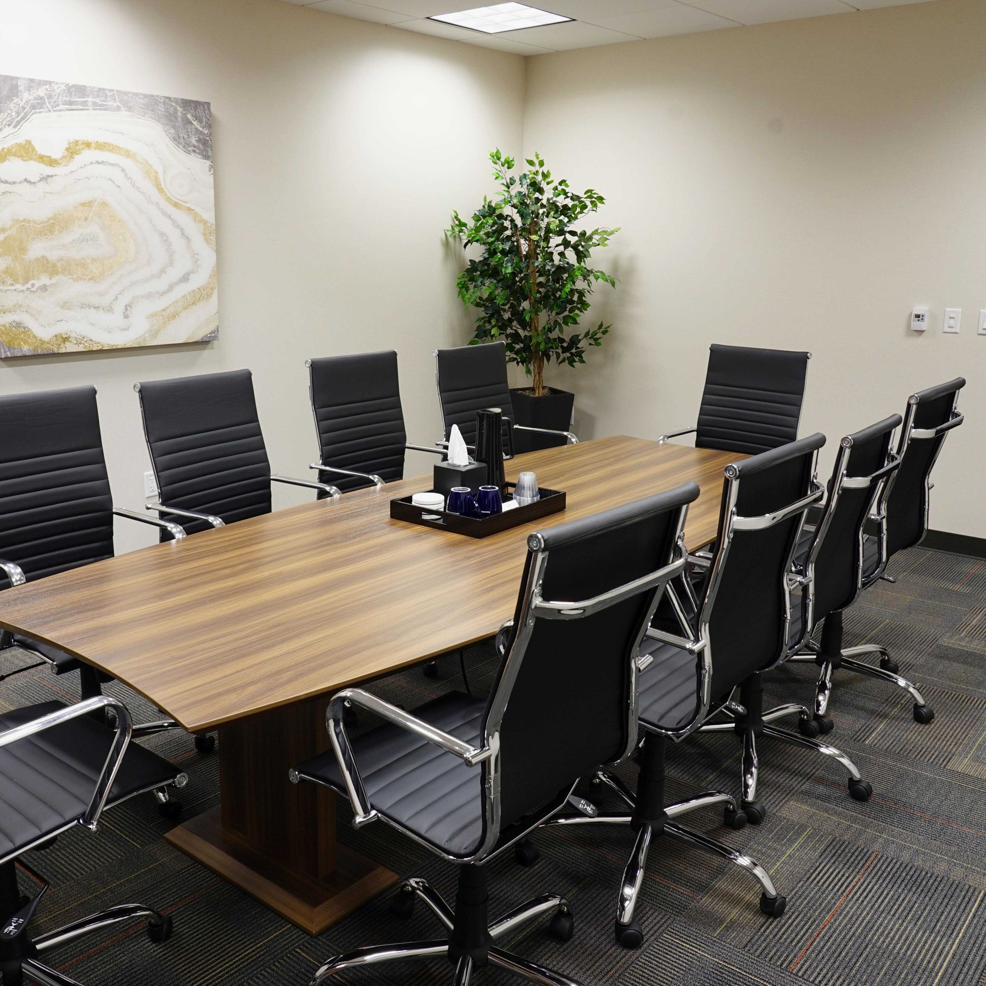 A modern conference room features a large wooden table surrounded by ten black leather chairs and a potted plant in the corner.