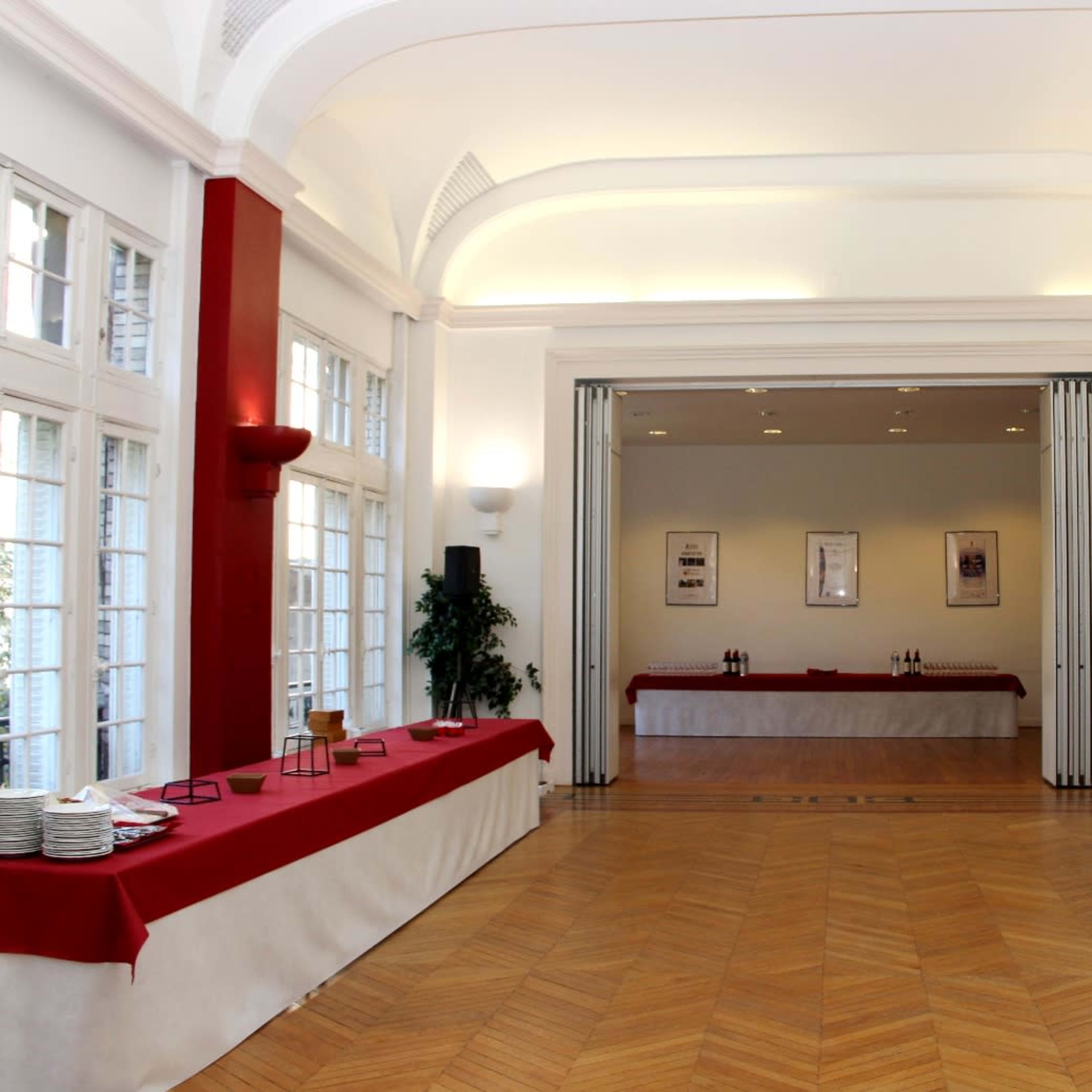 A spacious room with wooden flooring and white walls, featuring red table coverings along the sides and a set of sliding doors at the back.