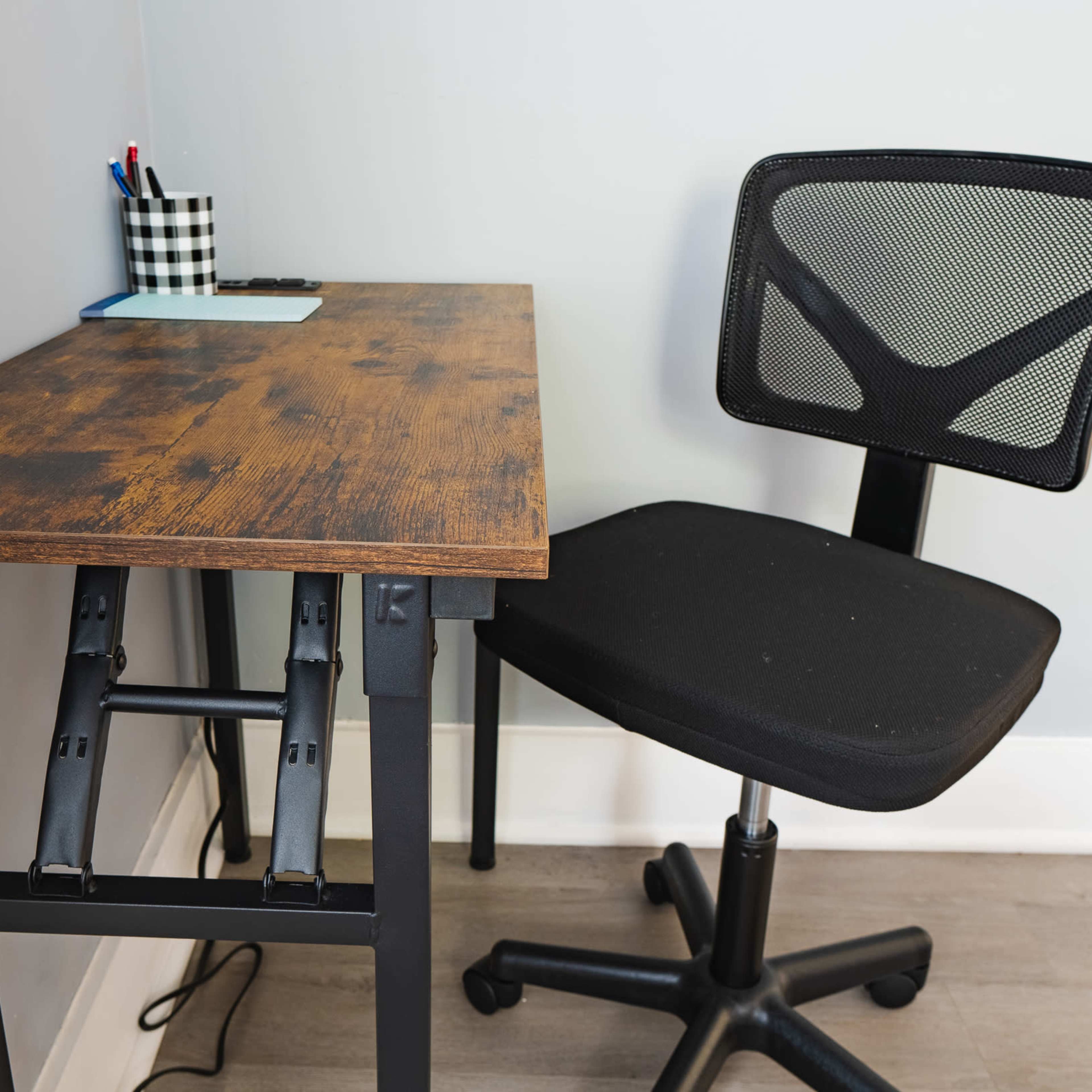 A wooden desk with a metal frame is positioned next to a black mesh office chair against a light gray wall.
