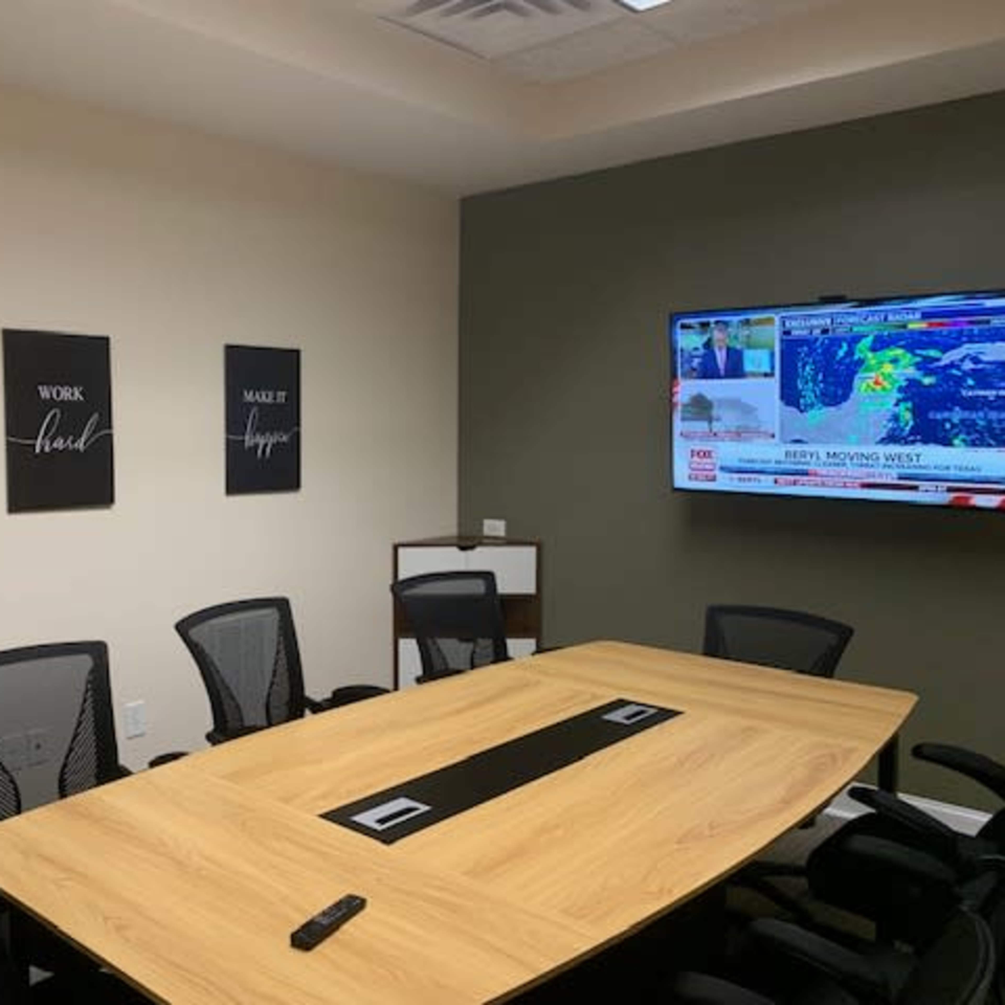 A conference room features a large wooden table surrounded by black chairs, with motivational posters on the wall and a television displaying a weather report.