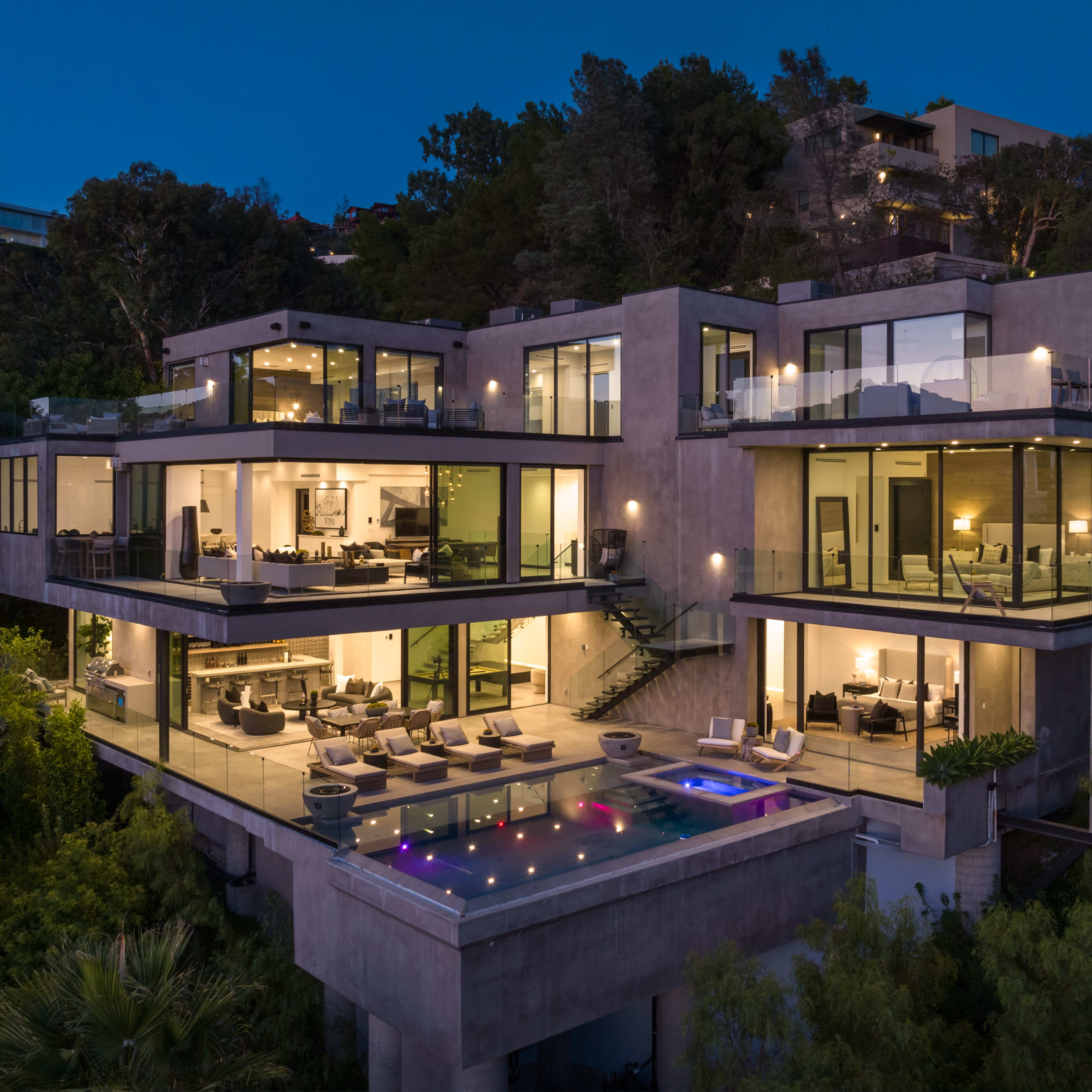 A modern multi-level house with large windows overlooks a pool area, surrounded by lush greenery and illuminated at night.