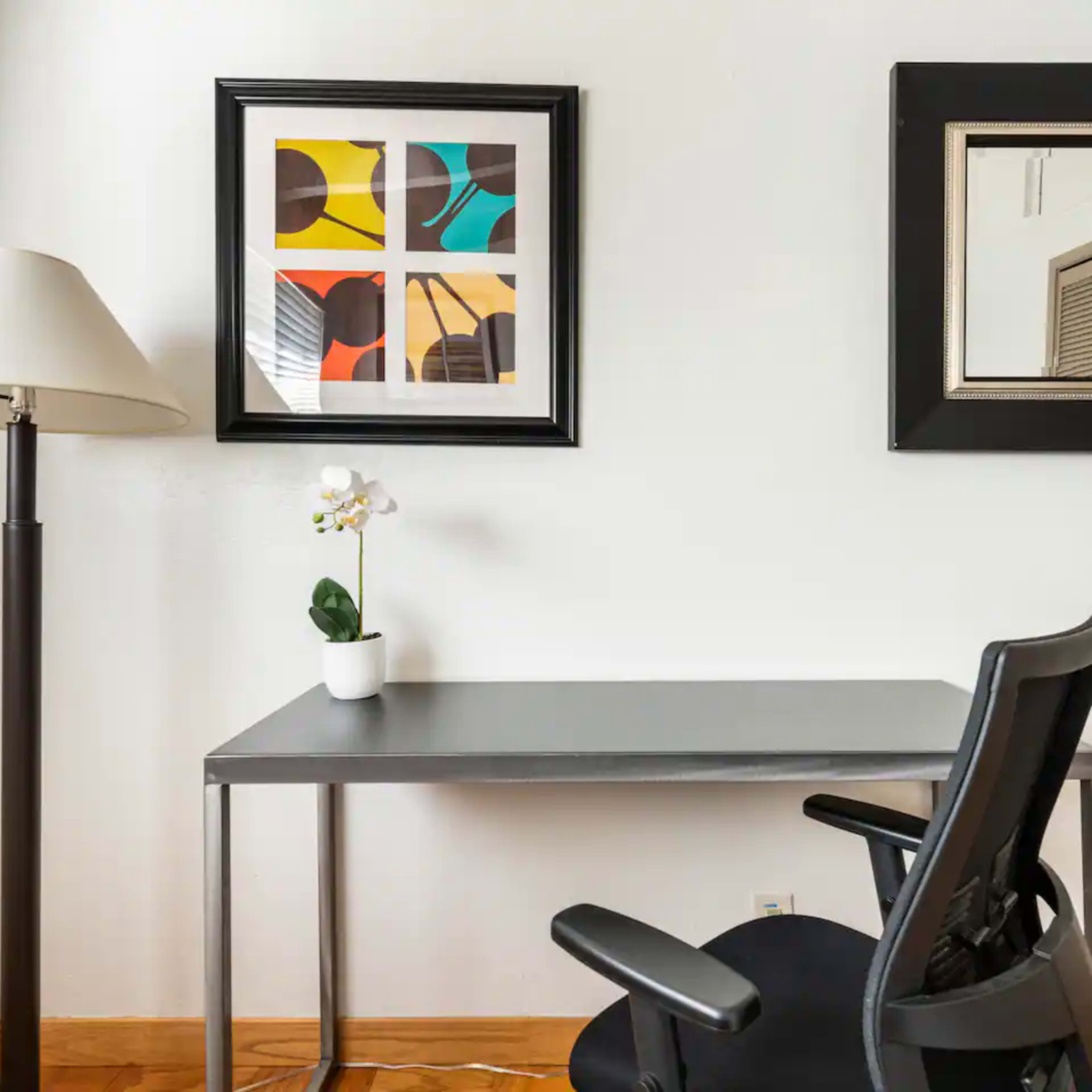 A modern workspace featuring a small desk, a chair, a lamp, a framed abstract artwork, and a mirror on the wall.