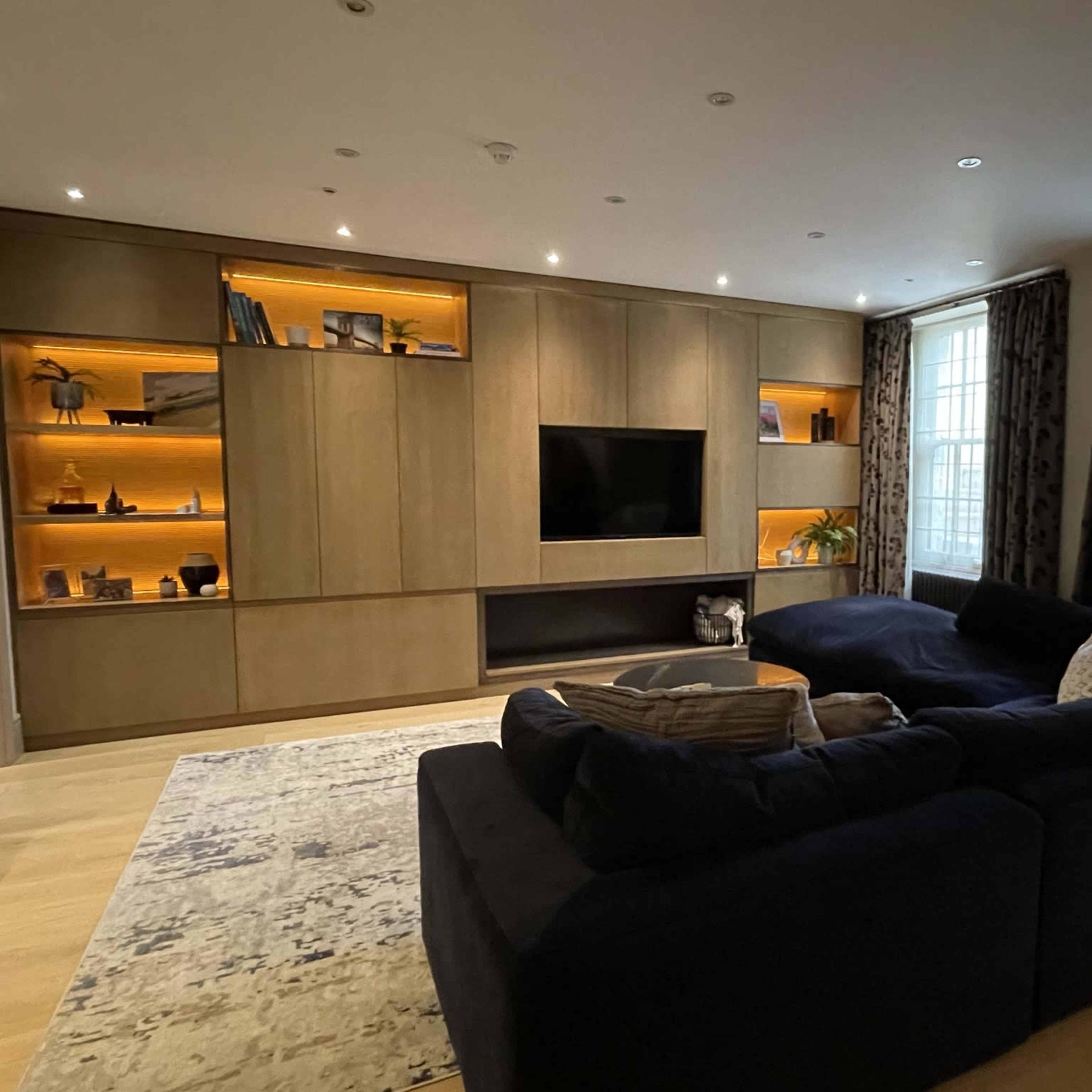 A modern living room with a dark sectional sofa, a large wall-mounted TV, and built-in shelves illuminated by warm lighting.
