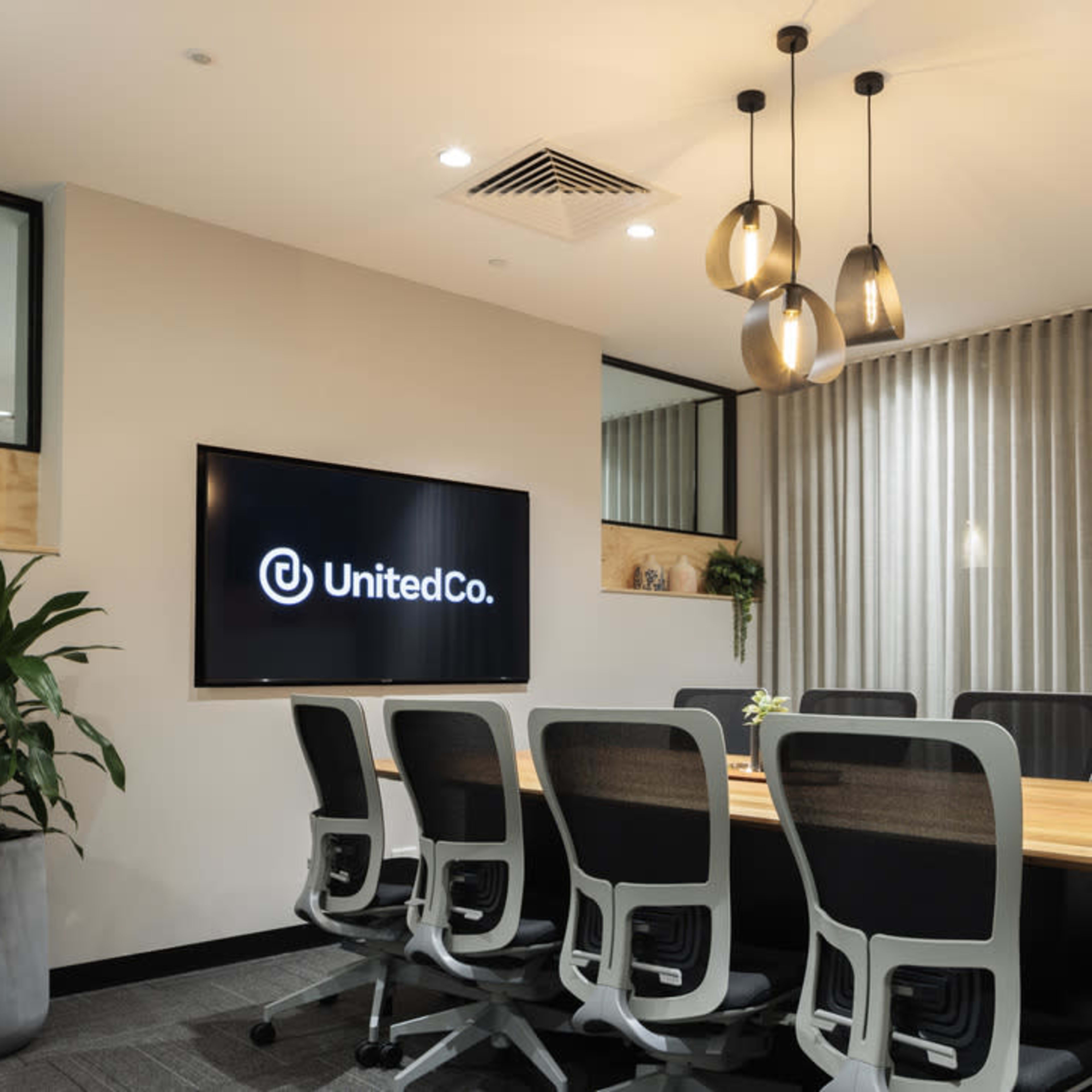A modern conference room features a long wooden table surrounded by ergonomic chairs, with a large screen displaying the UnitedCo logo on the wall.