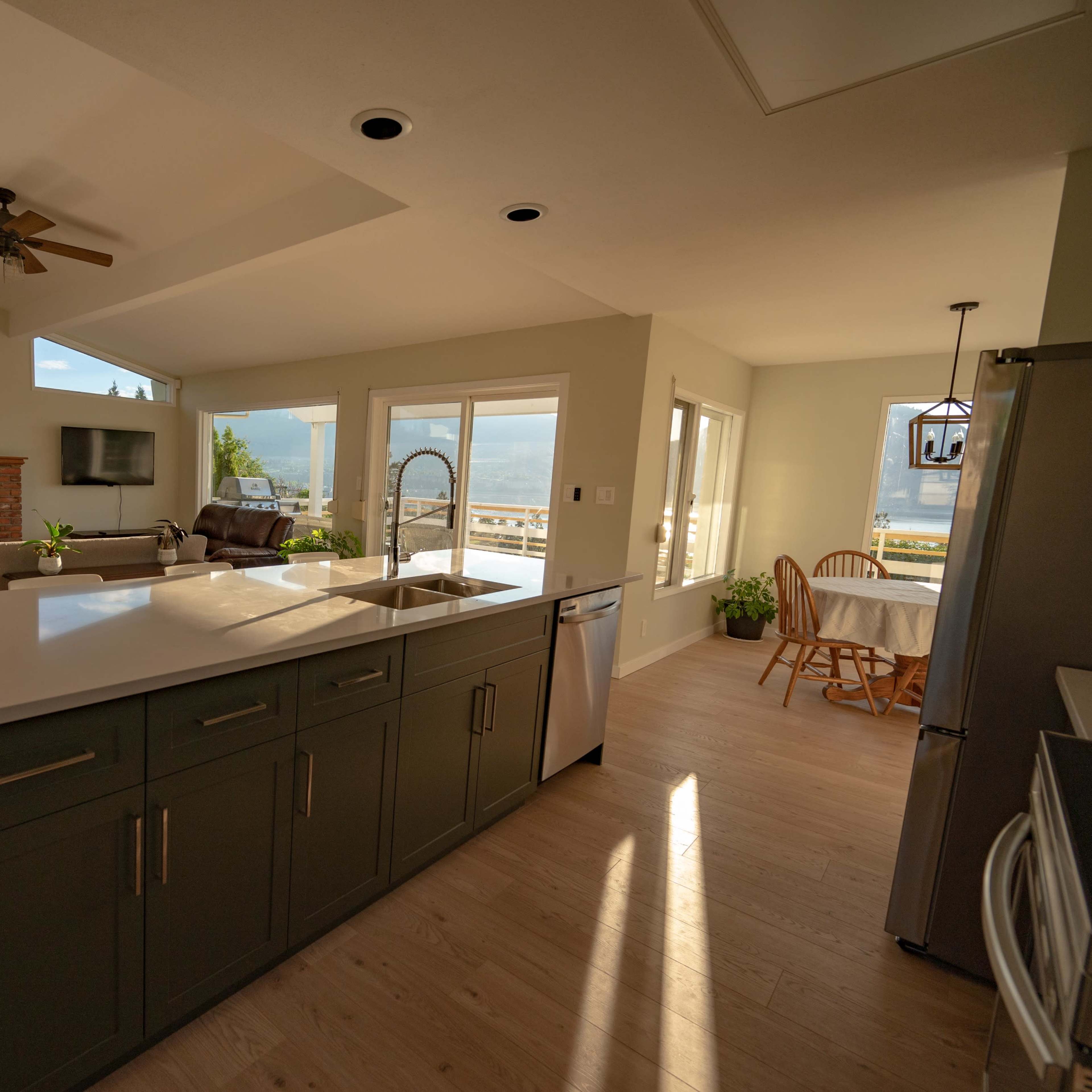 A brightly lit open-concept kitchen and living area with a stone fireplace, large windows, and a dining table near the kitchen.