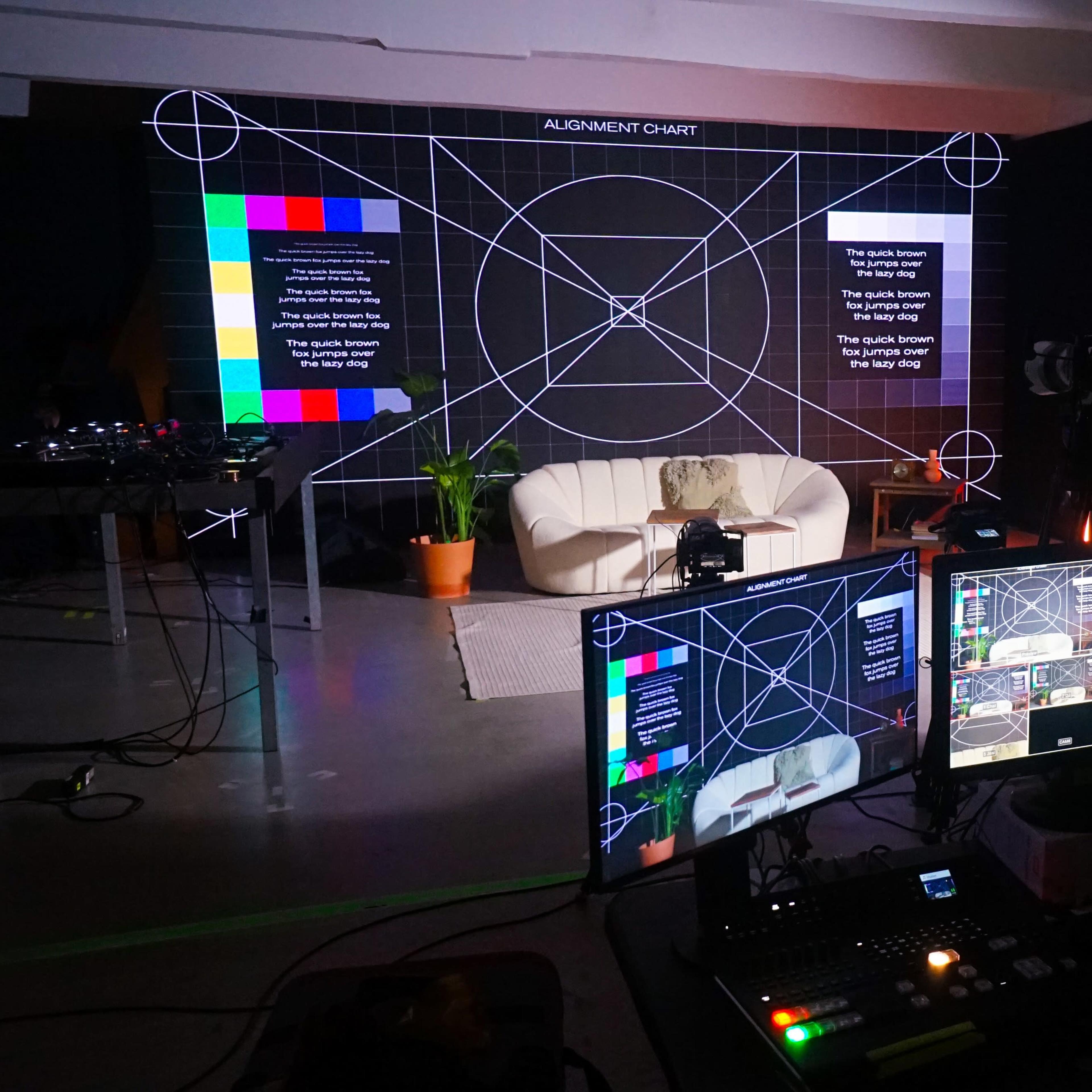 A modern studio setup featuring a white sofa, a large projection screen displaying an alignment chart, and multiple computer monitors used for broadcasting or video production.