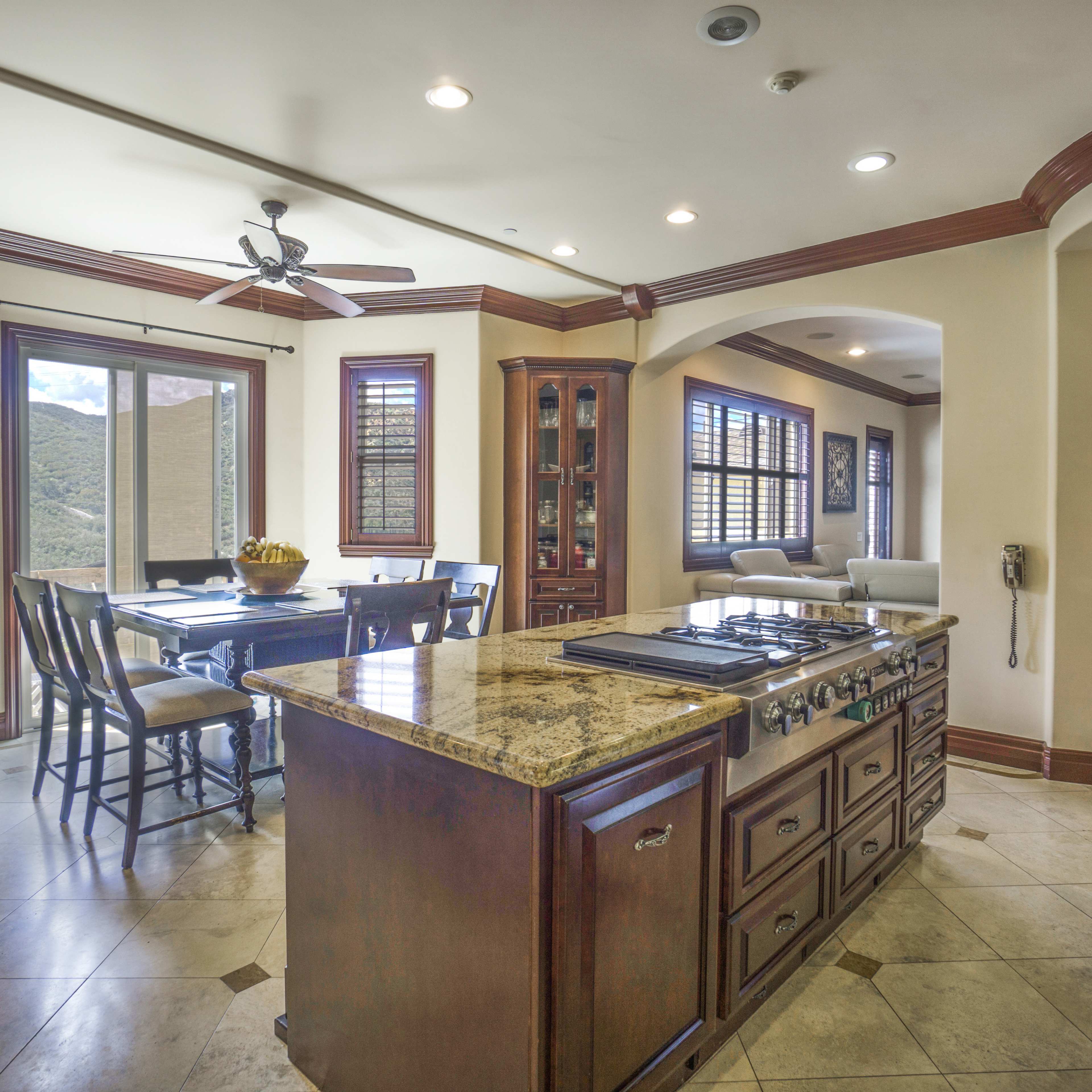 A spacious kitchen area featuring a central island with a gas stove, surrounded by wooden cabinetry and large windows that provide a view of the surrounding hills.