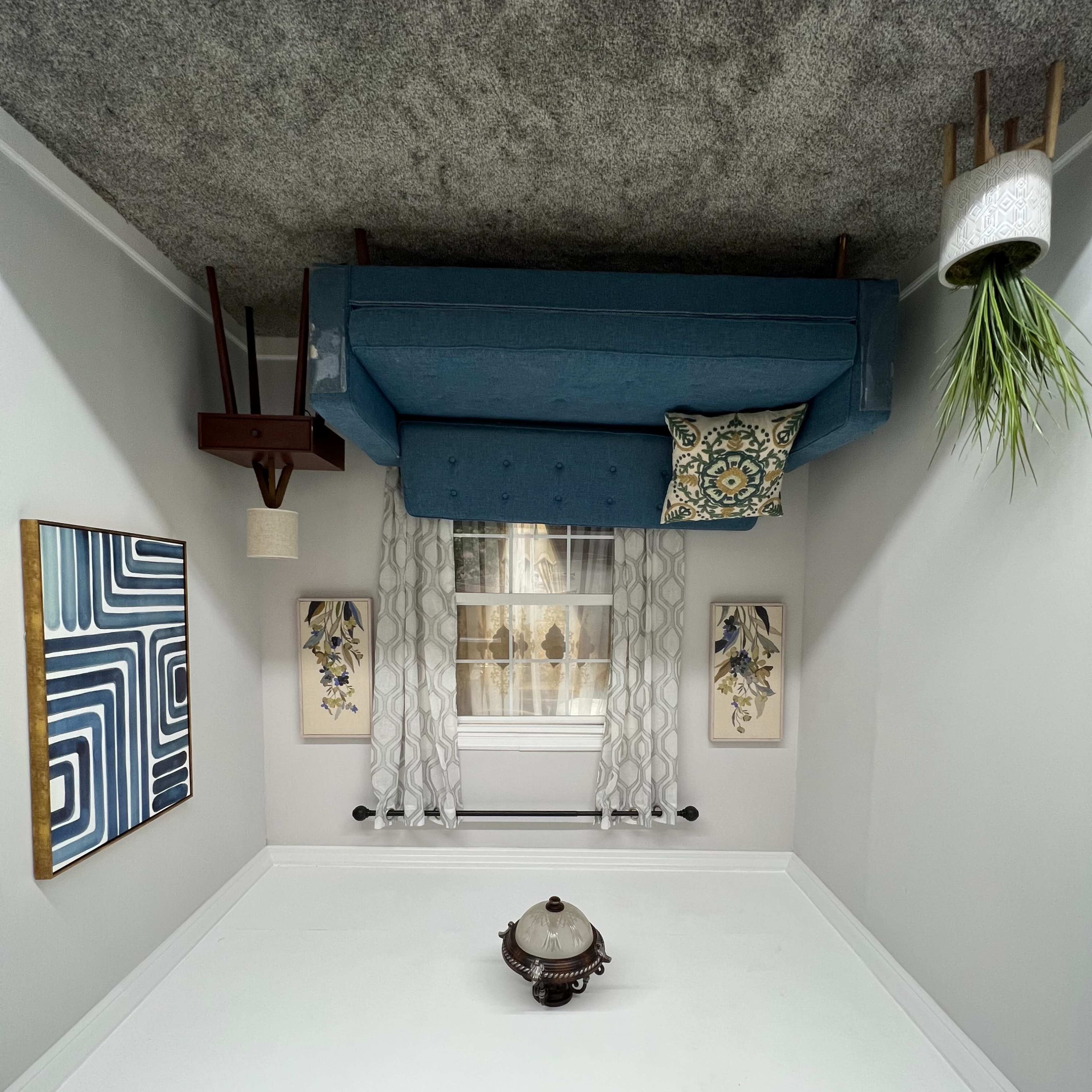 An inverted room with a blue couch mounted on the ceiling, accompanied by decorative wall art and a potted plant.