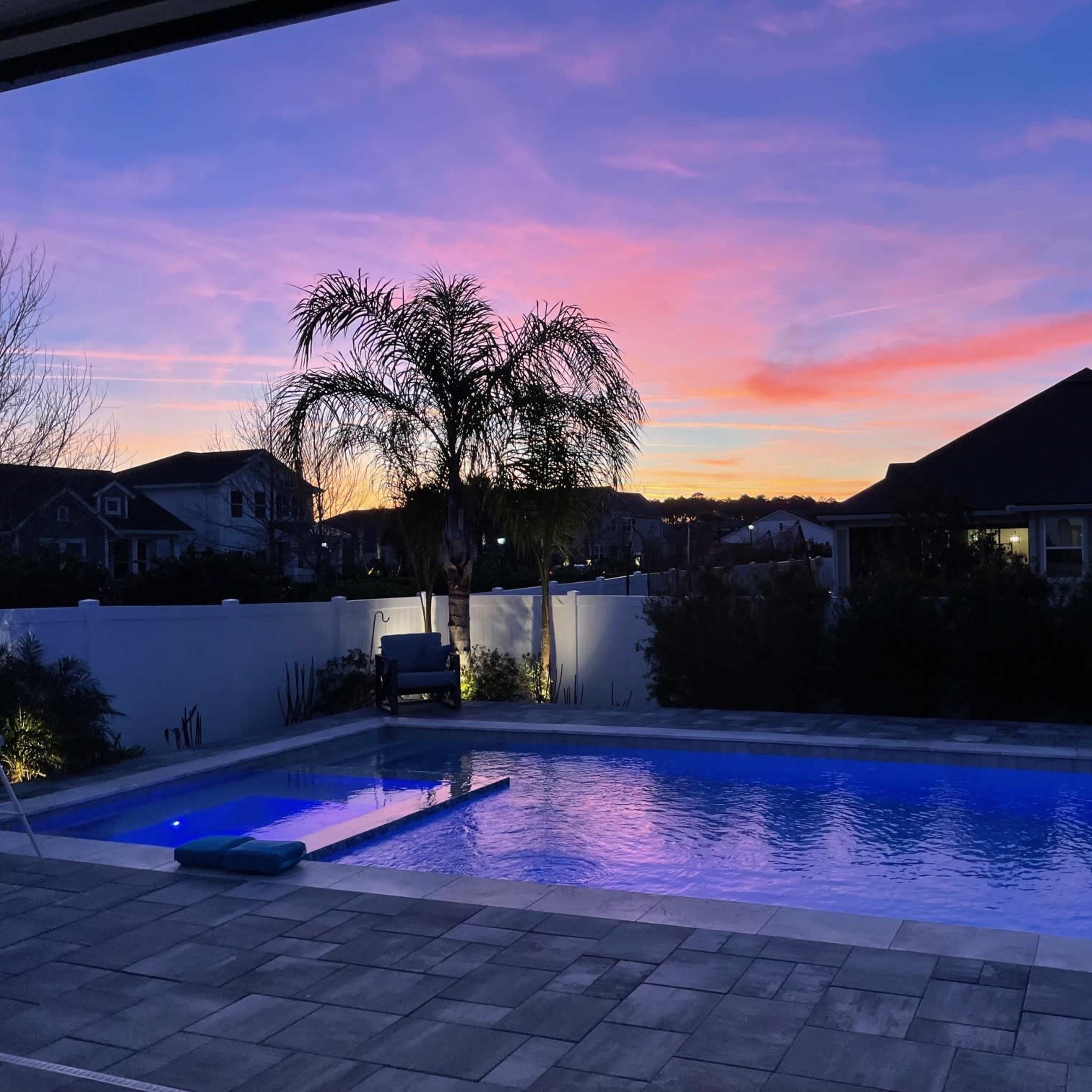 The scene shows a swimming pool illuminated by blue lights with a vibrant sunset sky and houses in the background.