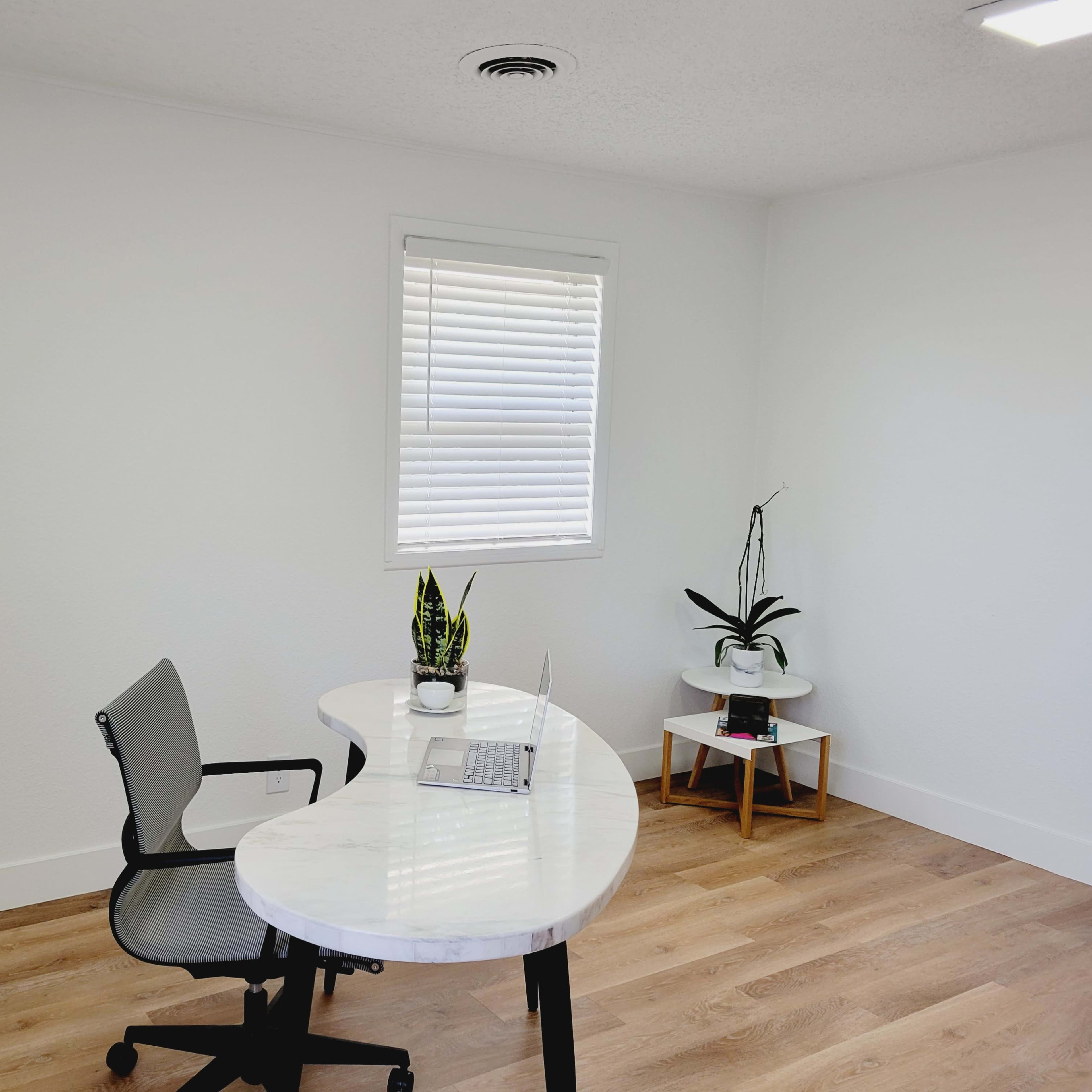 An office space features a white desk with a laptop, a chair, and potted plants next to a window.