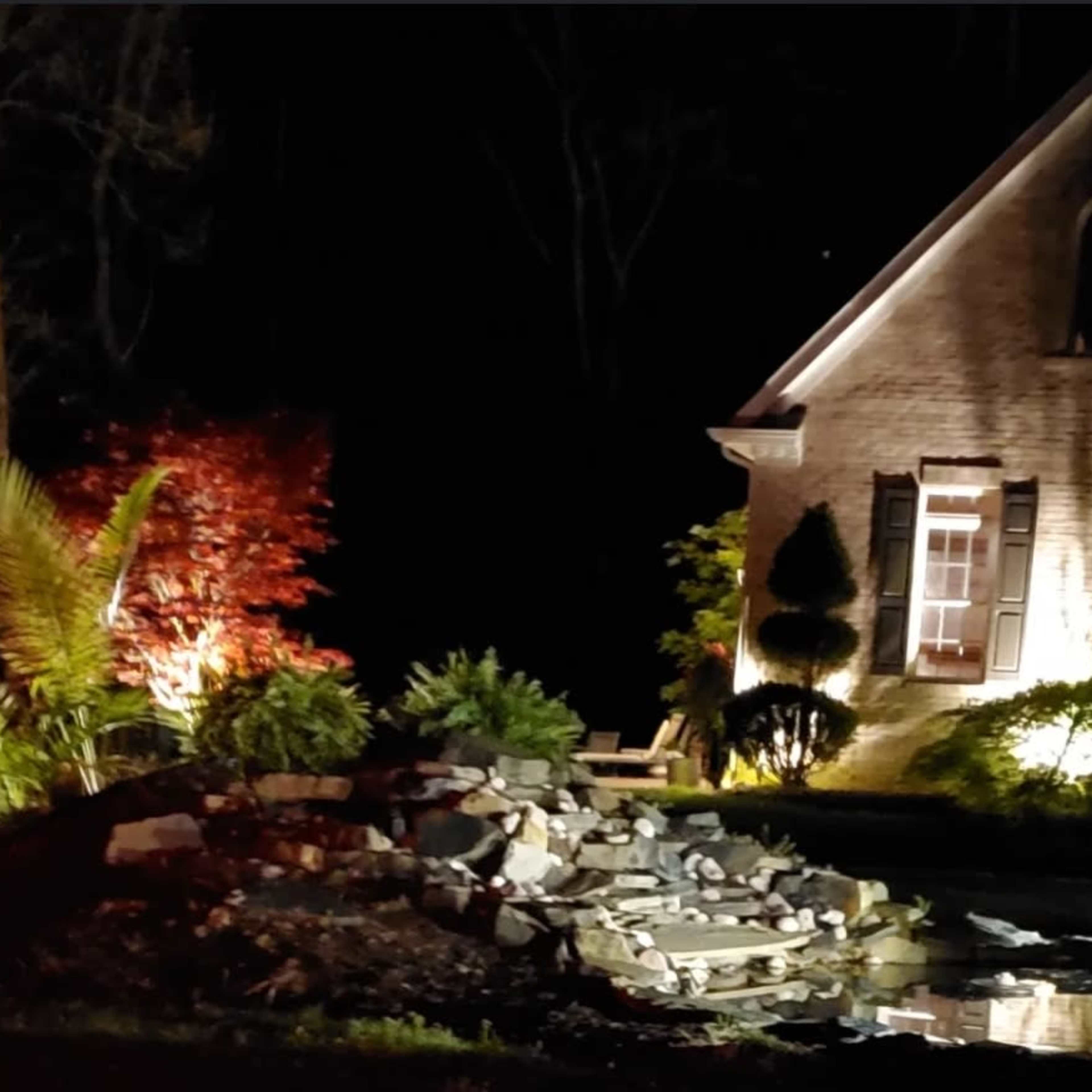 A lit brick house stands next to a landscaped area featuring a pile of rocks and a small pond, surrounded by plants.