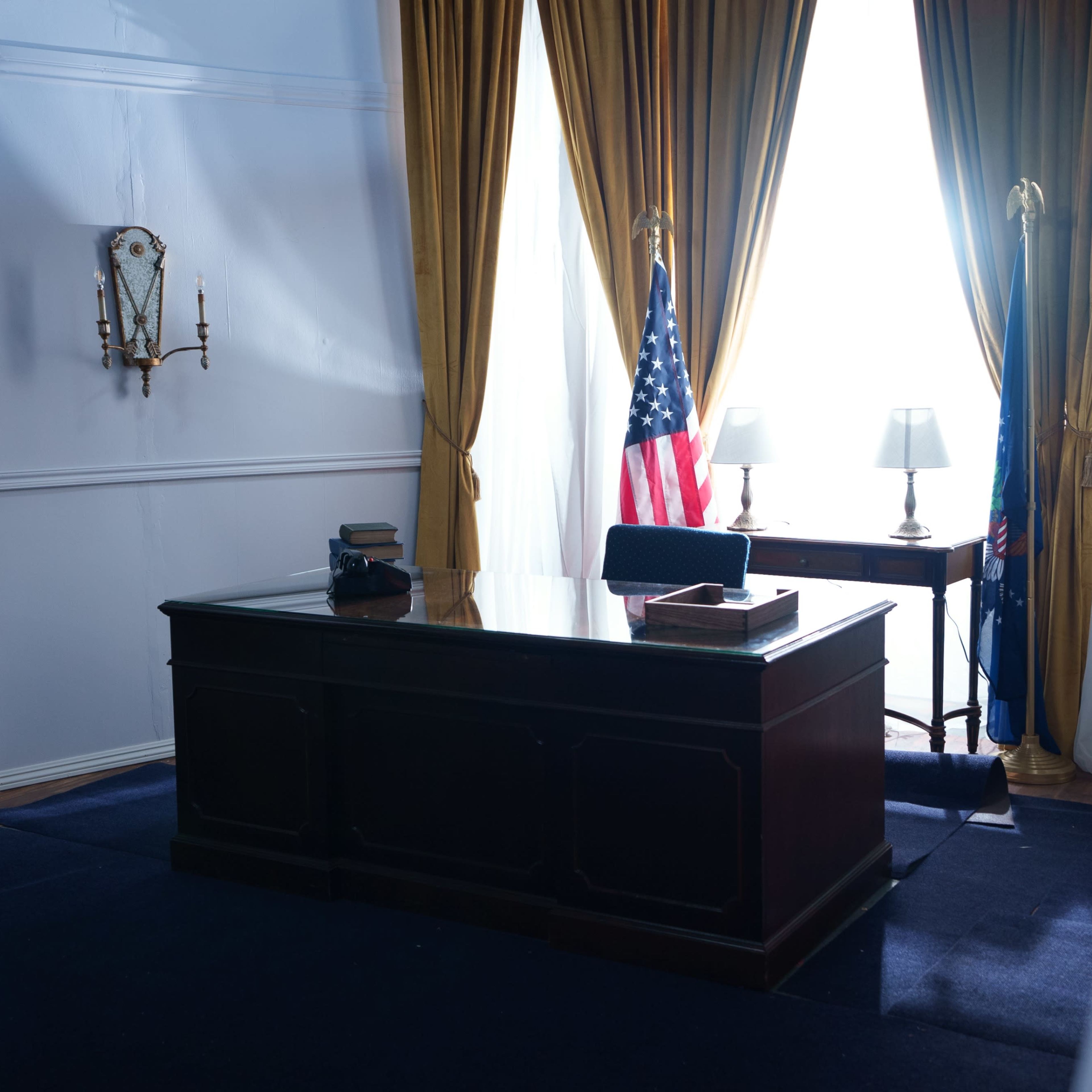 A formal office space with a wooden desk, two lamps, an American flag, and large windows draped with yellow curtains.