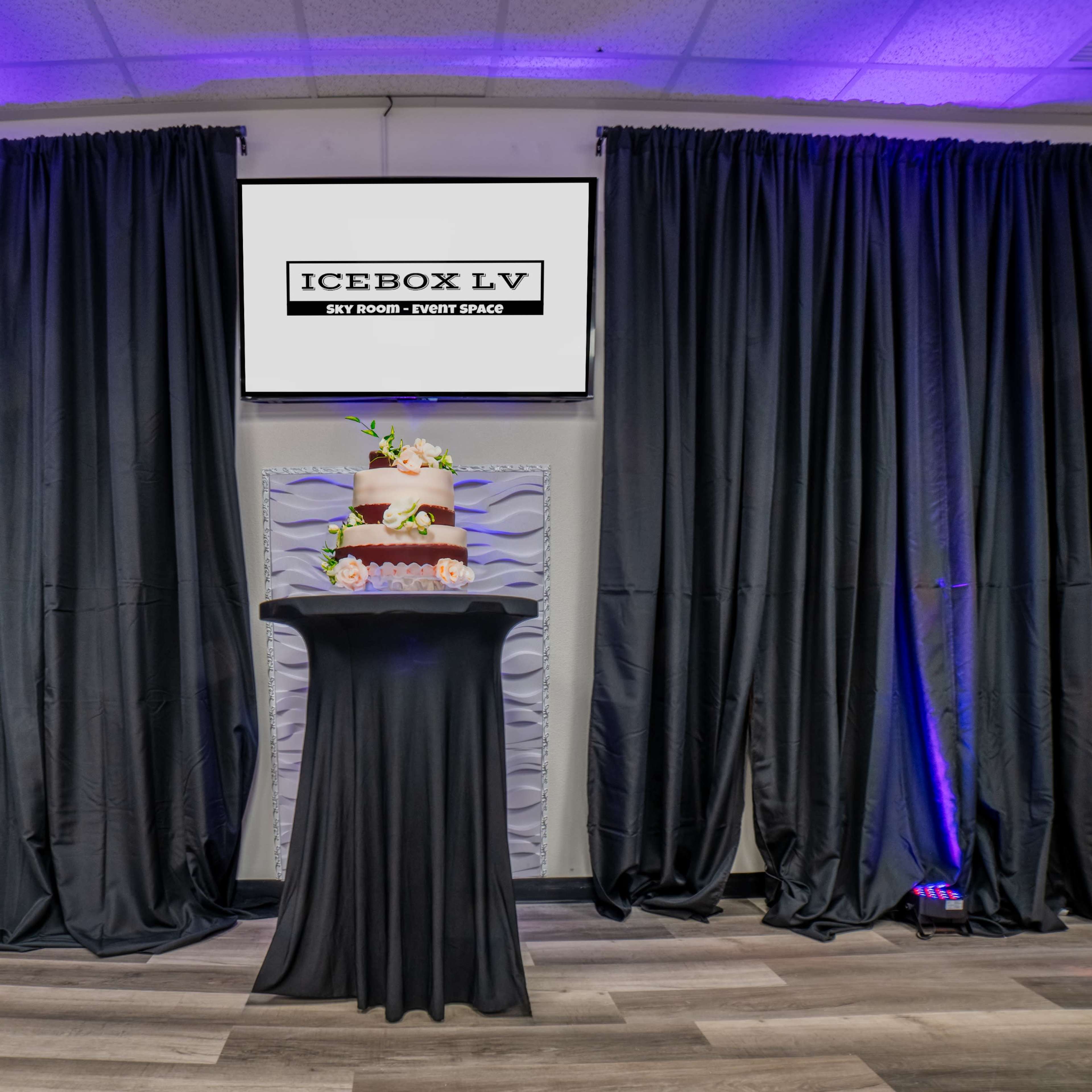 A decorated cake is displayed on a pedestal in front of a large screen and black curtains in an event space.