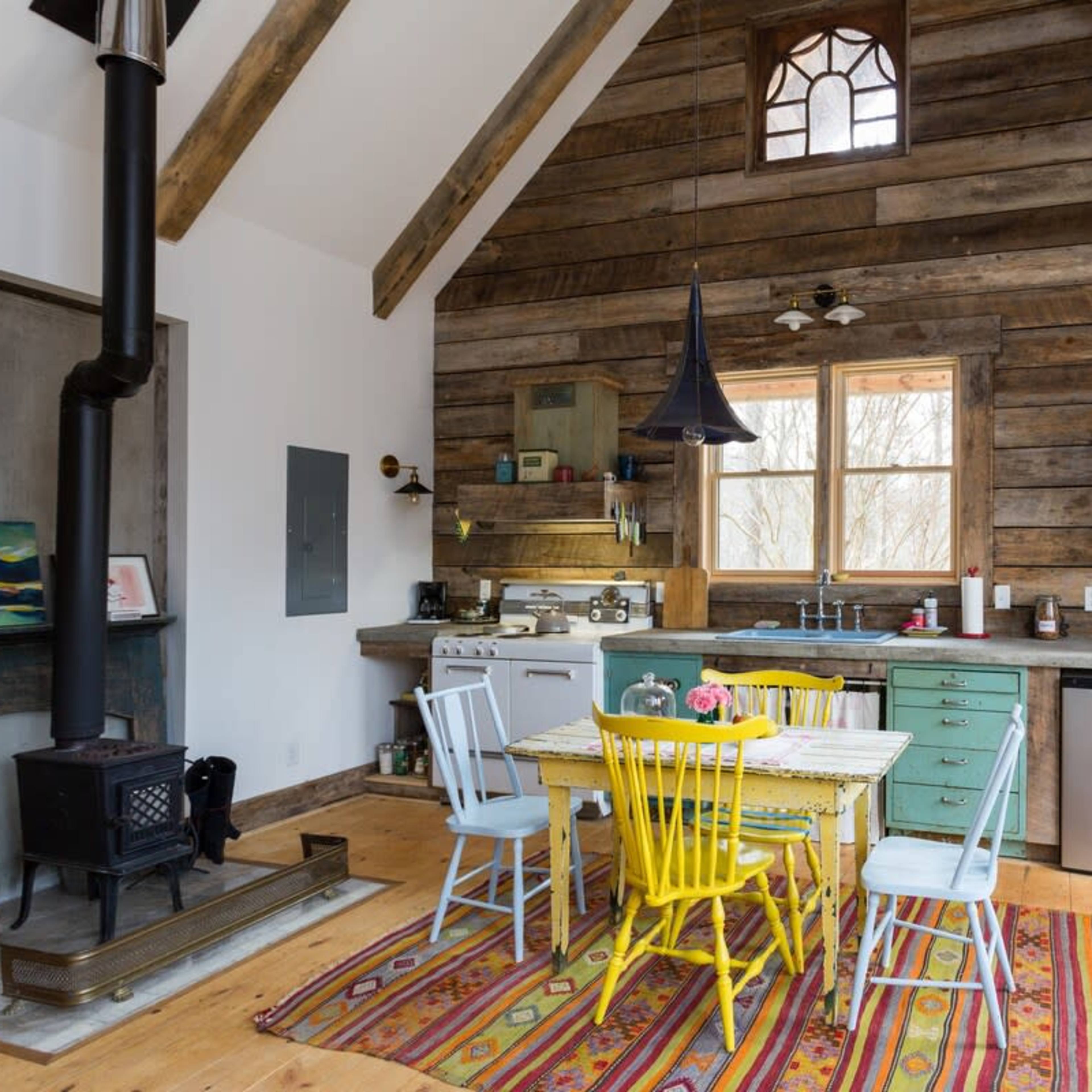 A rustic kitchen features a wooden dining table with colorful chairs, a stove, and a large window overlooking the outdoors.