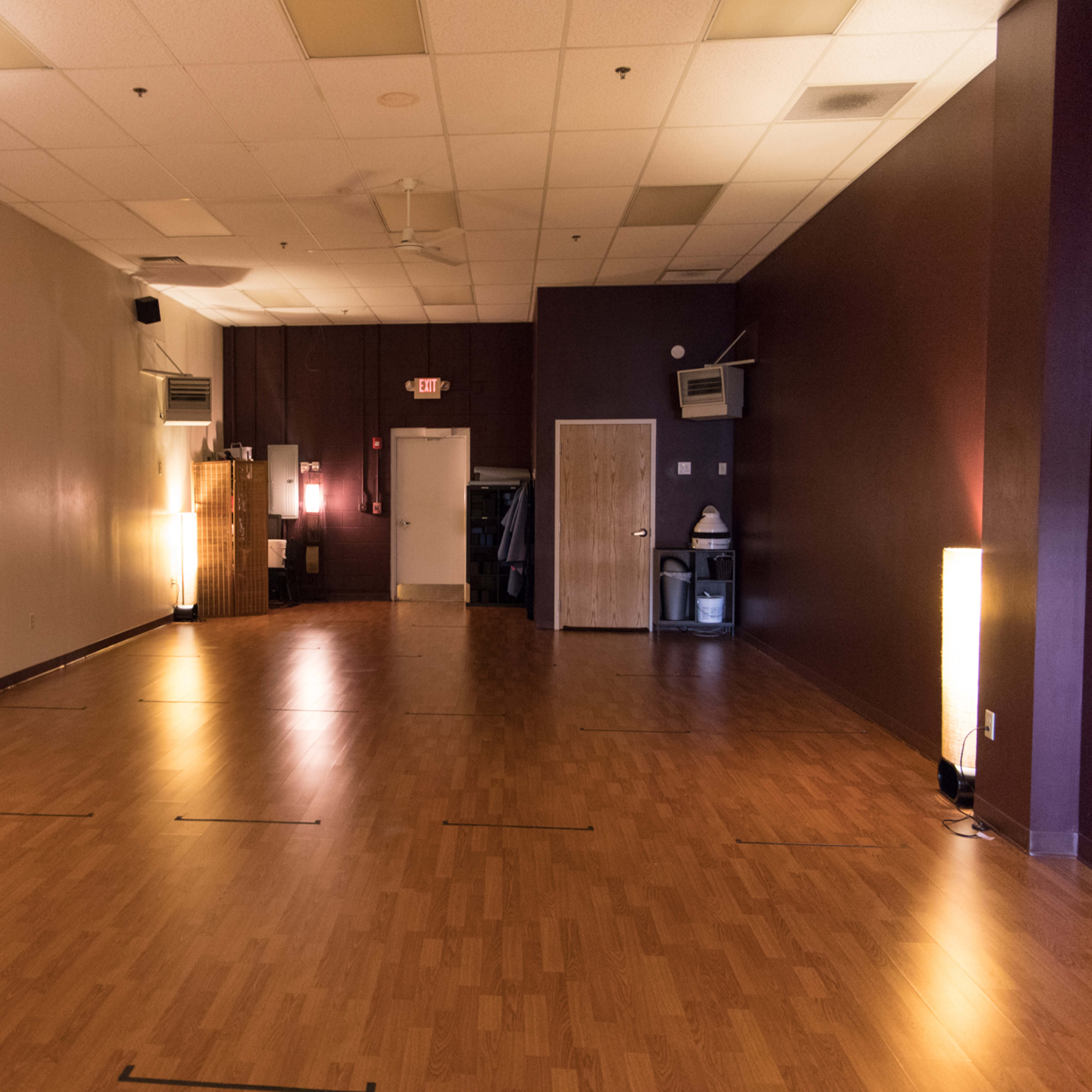 An empty, well-lit studio space with wooden flooring, featuring two tall lamps on either side and a door leading to another room.