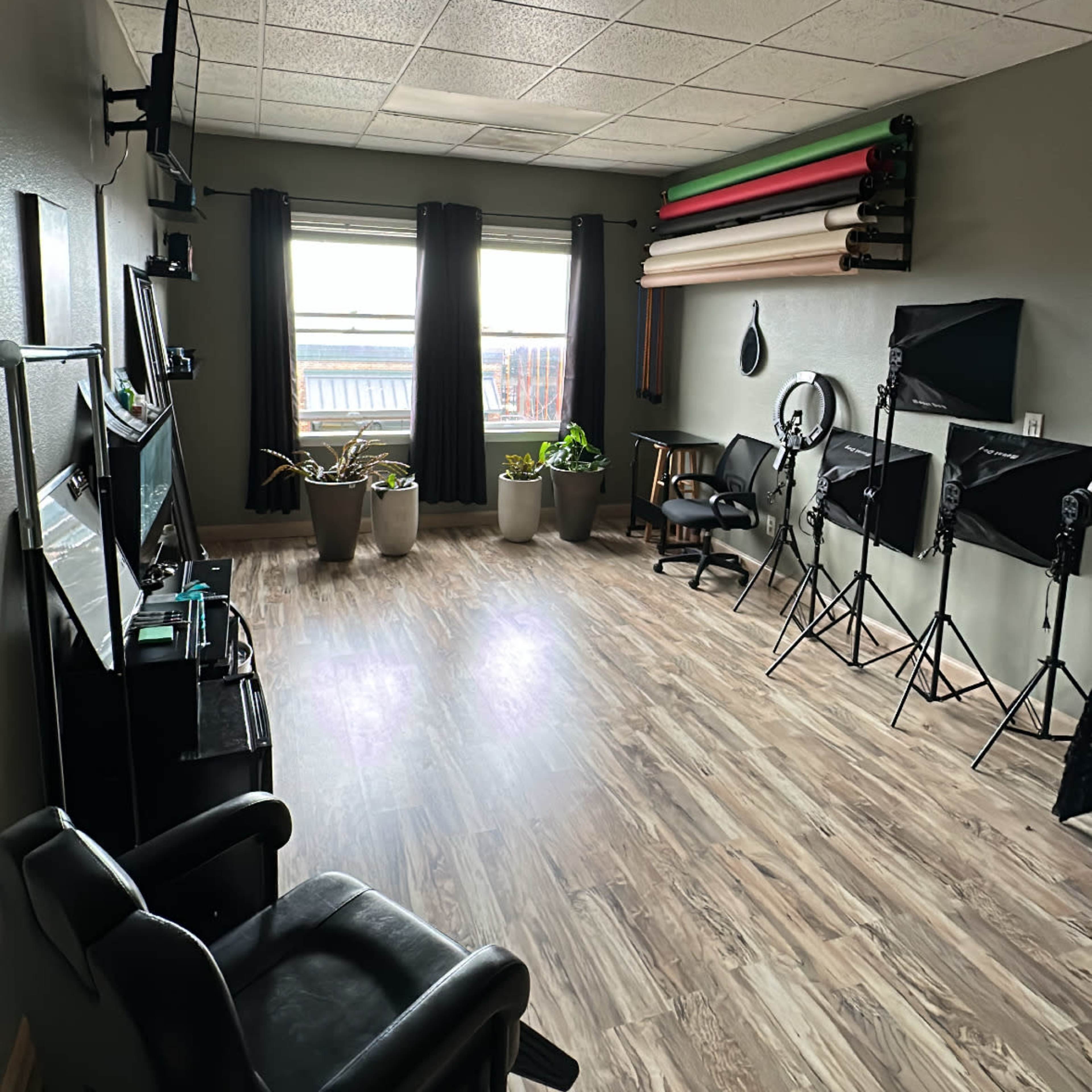 A spacious studio with wooden flooring, two large windows, several potted plants, and equipment for photography or videography, including light stands and colorful backdrops mounted on the wall.
