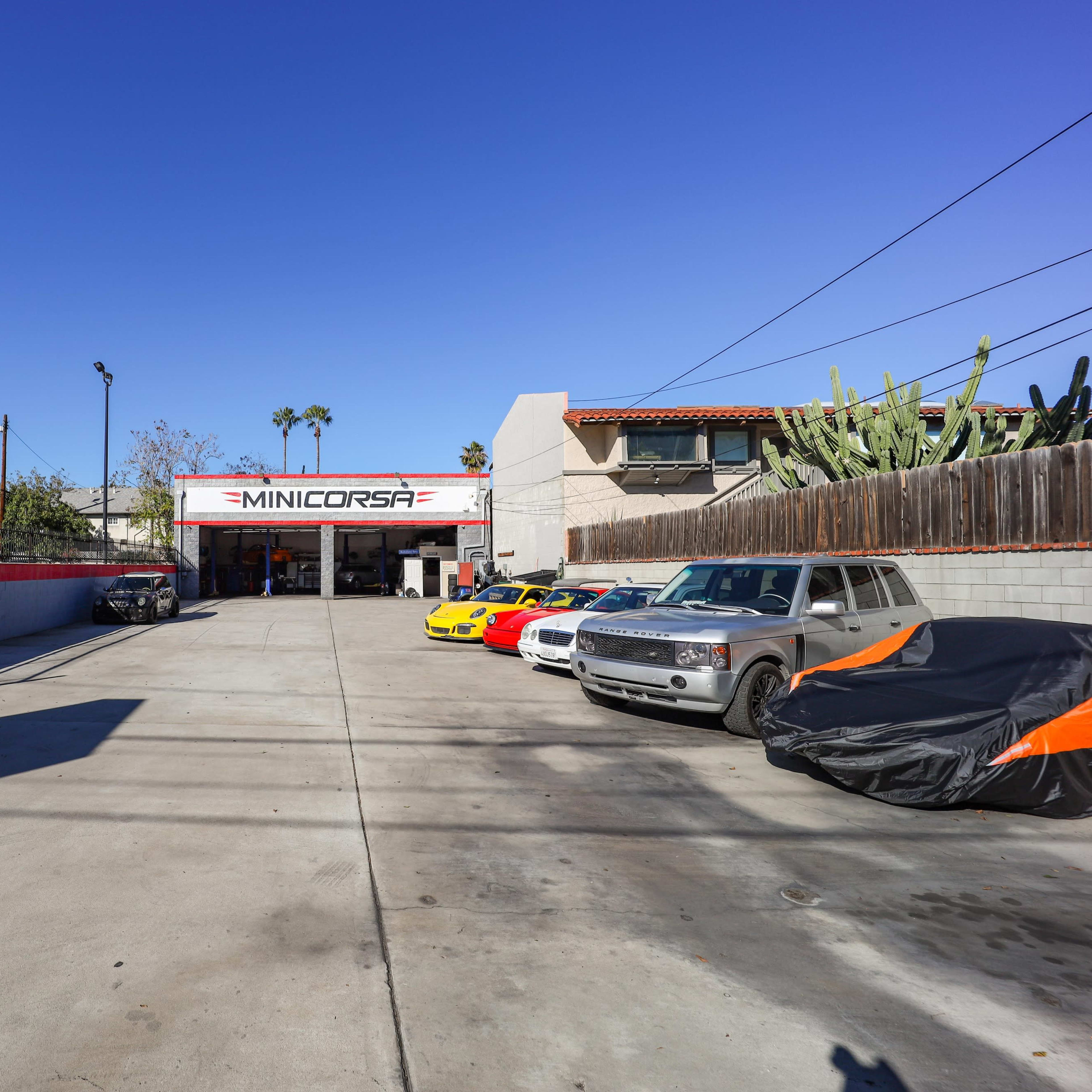 A clear view of a car workshop named "MINICORSA," featuring several cars parked in front and a bright blue sky overhead.