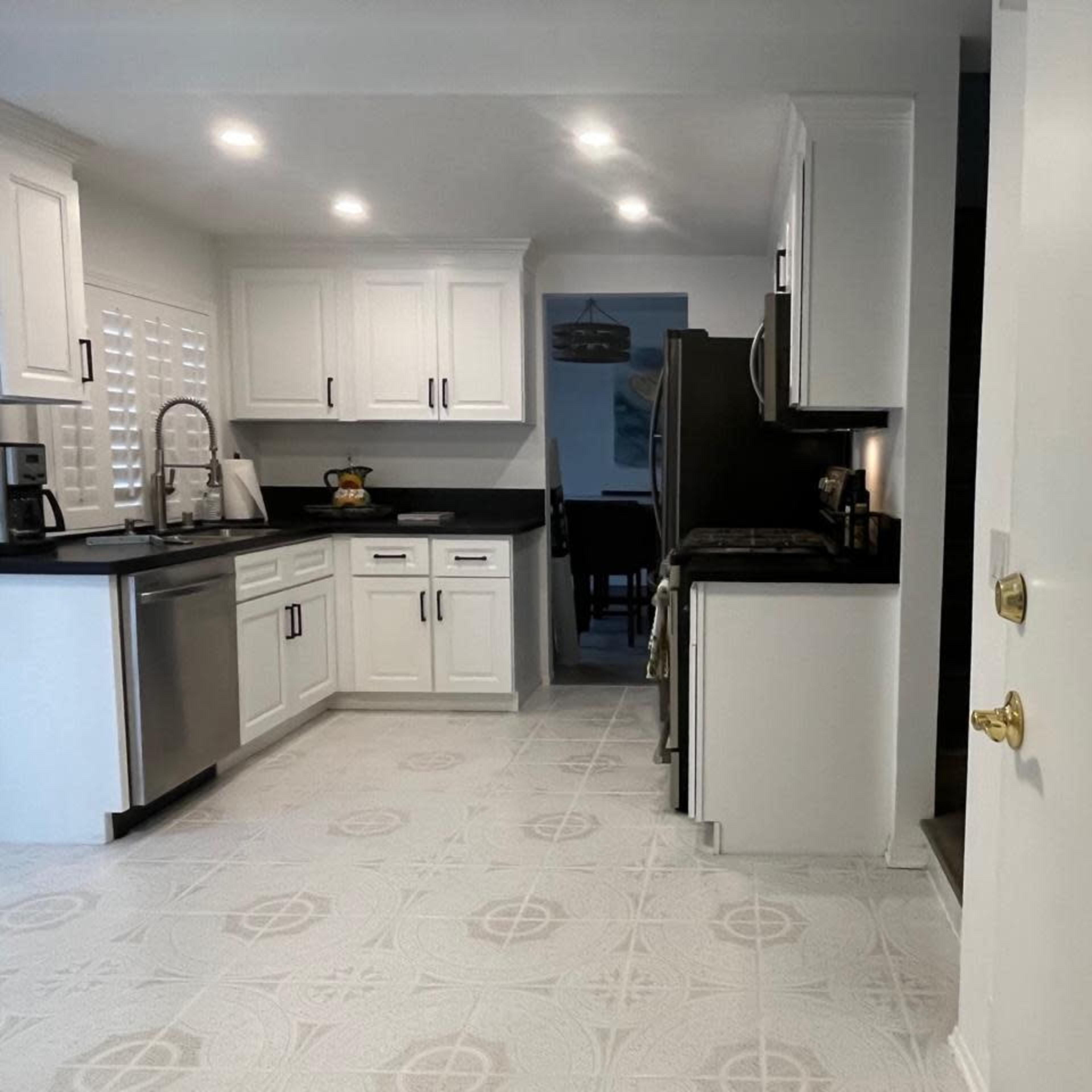 A modern kitchen with white cabinetry, black countertops, and a tiled floor, leading to a doorway at the far end.