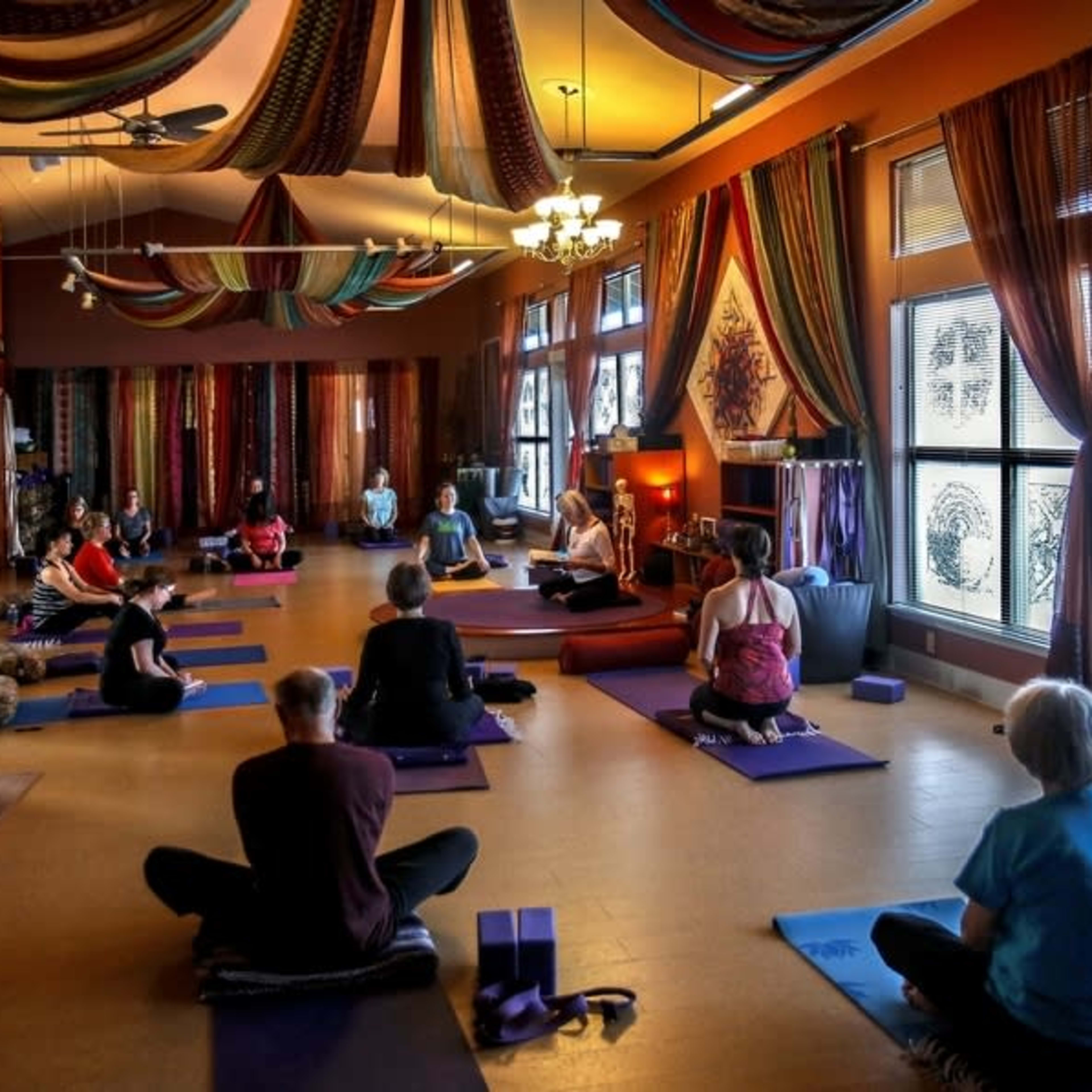 A group of people participates in a yoga class in a warmly decorated studio, seated on mats and facing an instructor at the front.