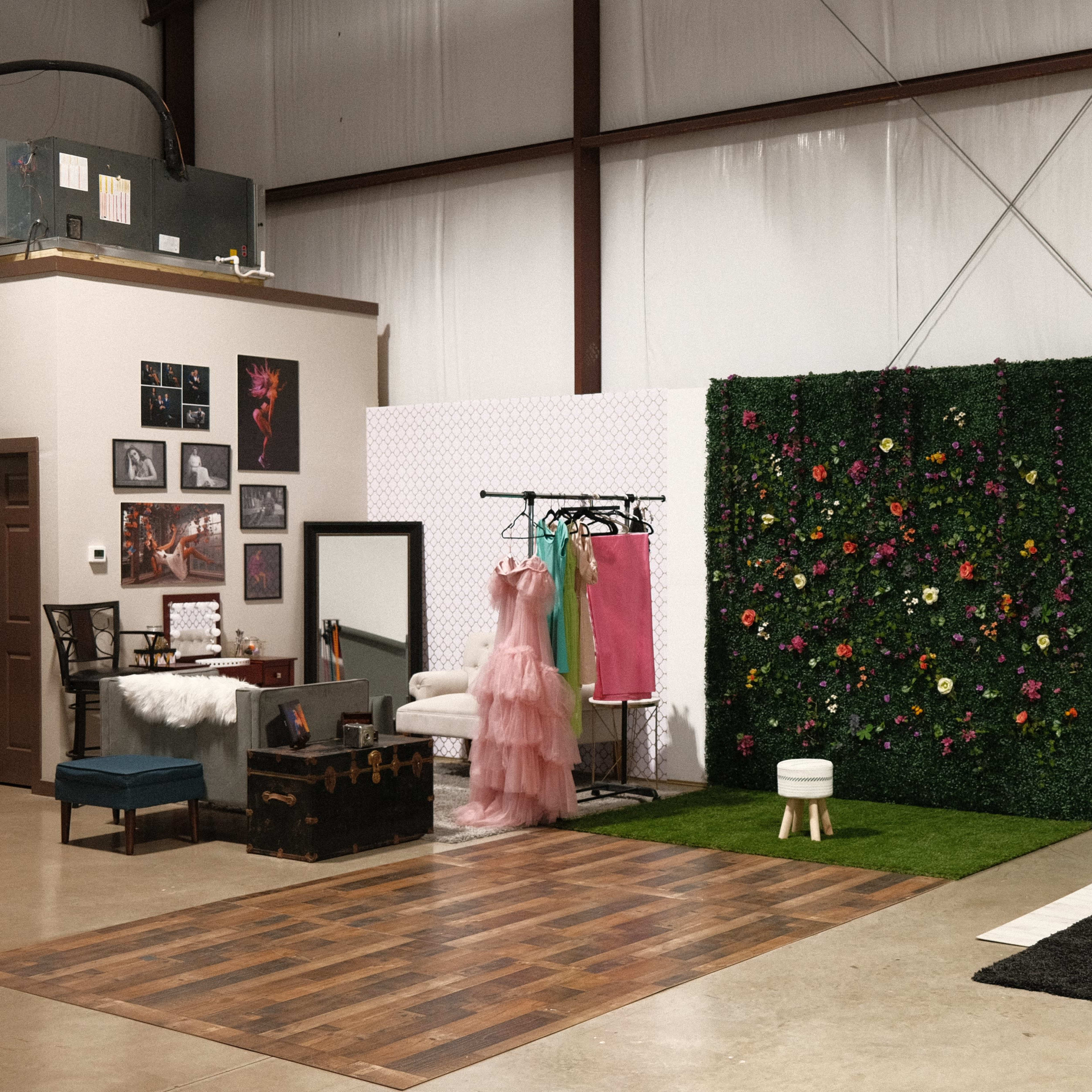 A studio space featuring a mix of furniture including a couch, a dressing area with a mirror, clothing racks, and a decorative wall with greenery and art.
