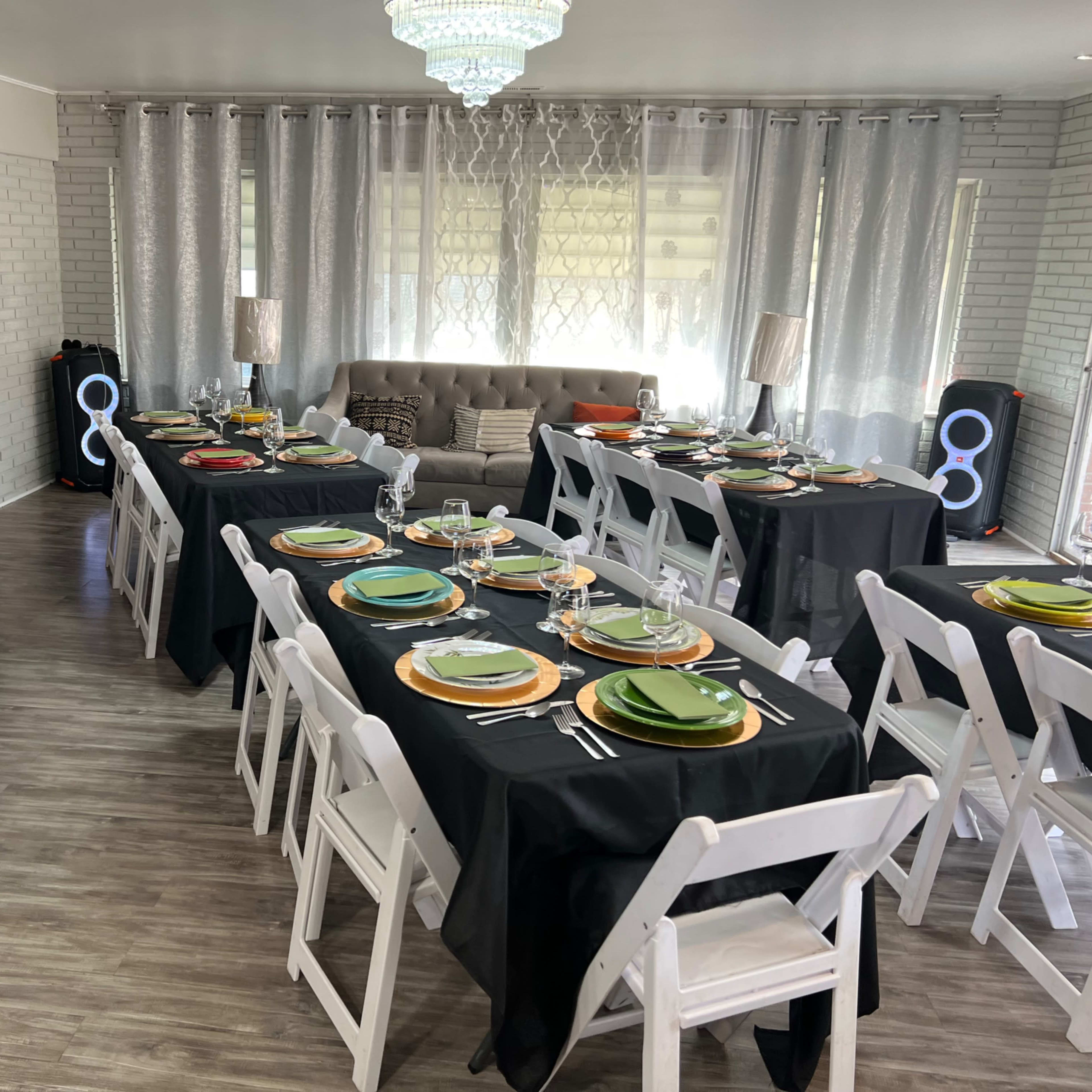 A spacious dining area is set up with multiple tables covered in black cloths, each arranged with colorful plates, utensils, and glasses, alongside a plush sofa and decorative lighting.