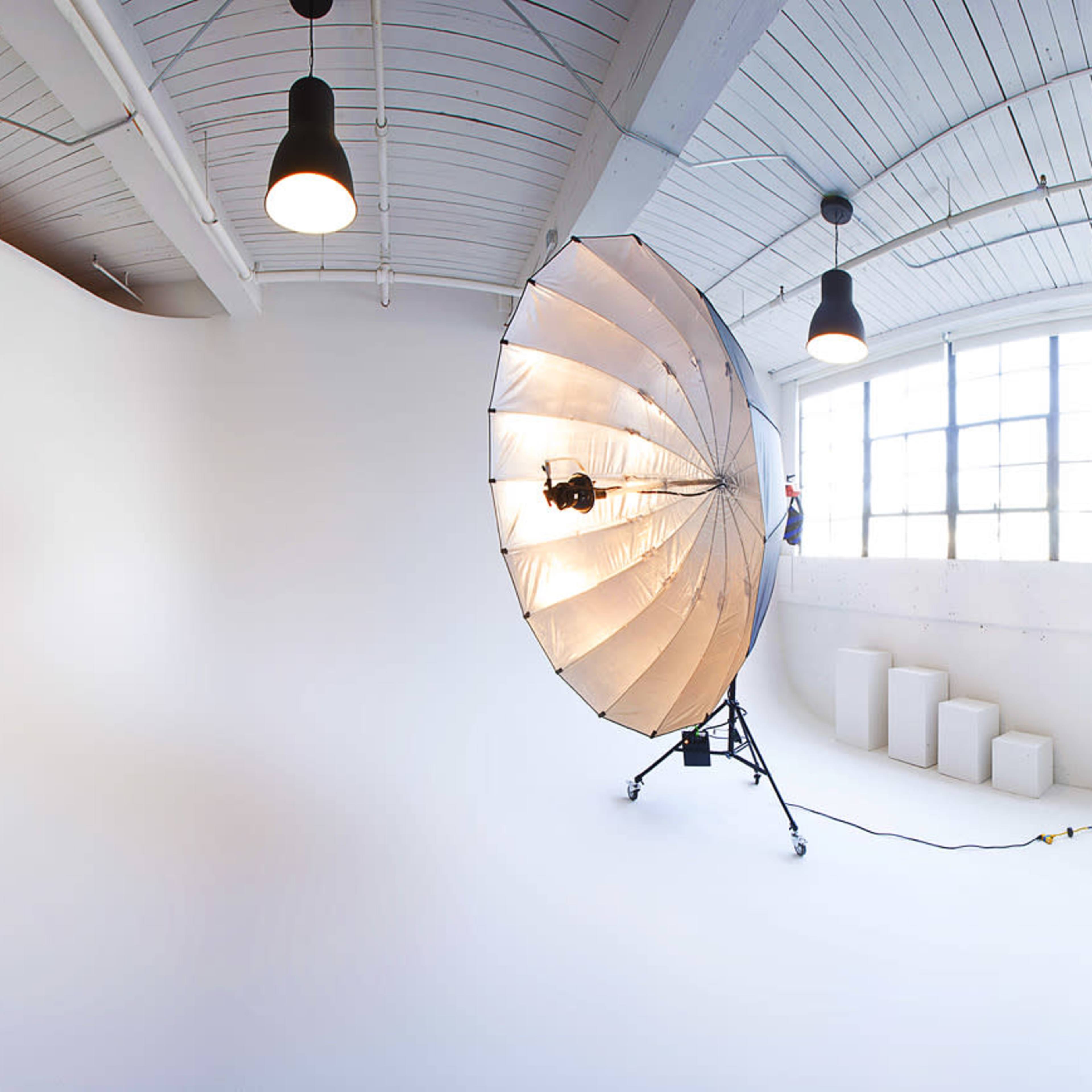 A spacious photography studio with a large umbrella light, a table, seating area, and large windows.