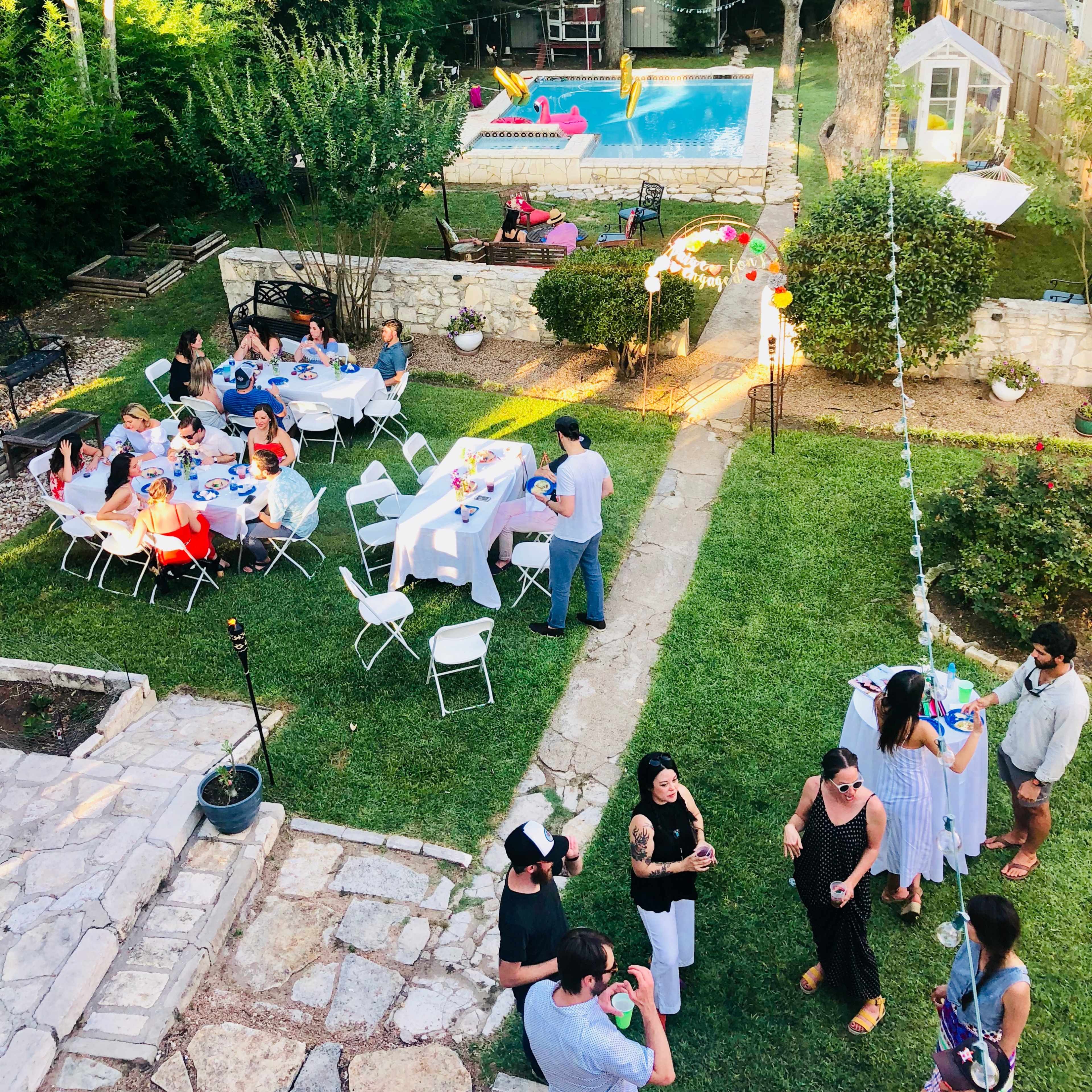 The backyard features a pool surrounded by tables and chairs, with people gathered for an outdoor event.