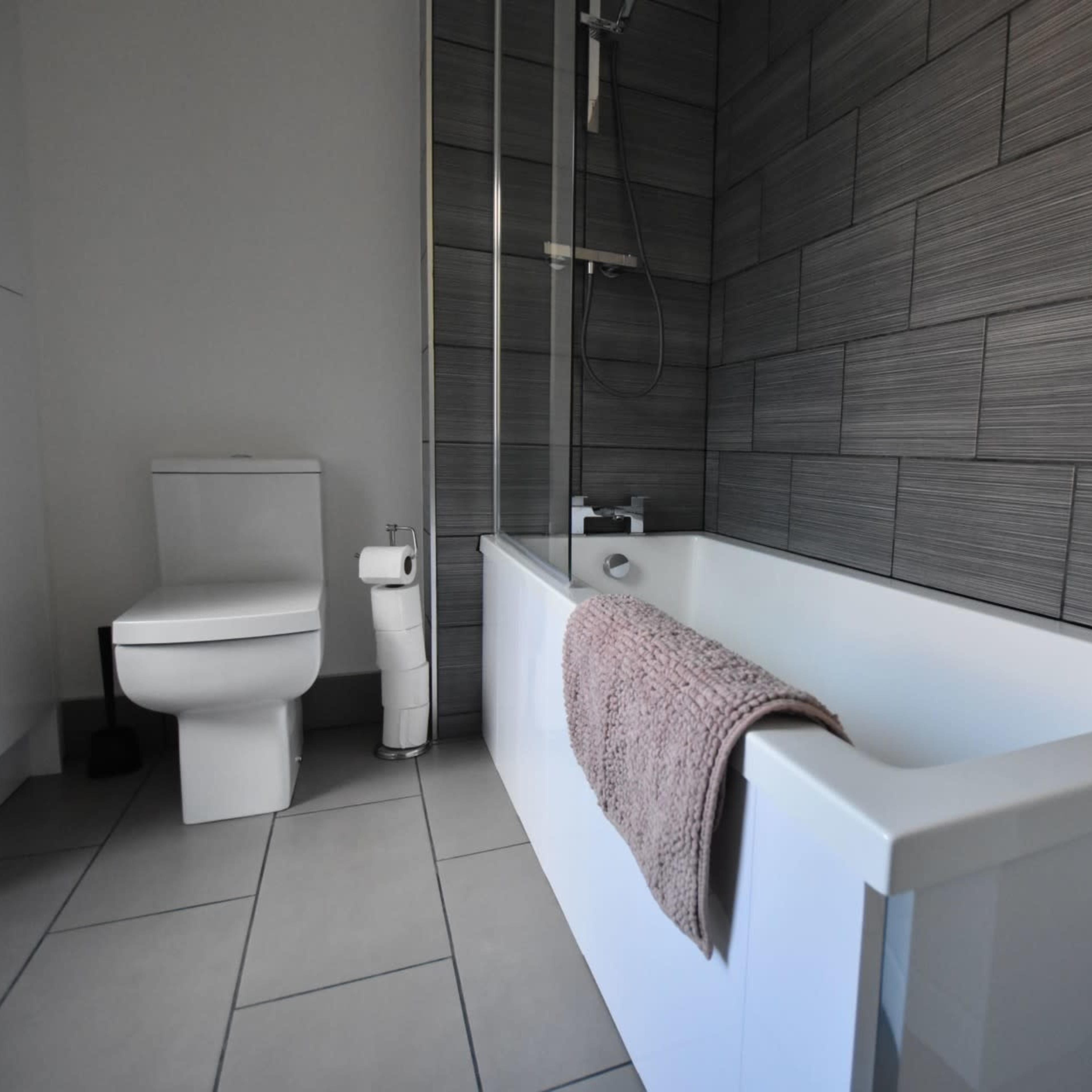 A modern bathroom featuring a white bathtub with a towel draped over it, a toilet, and grey tiled walls.